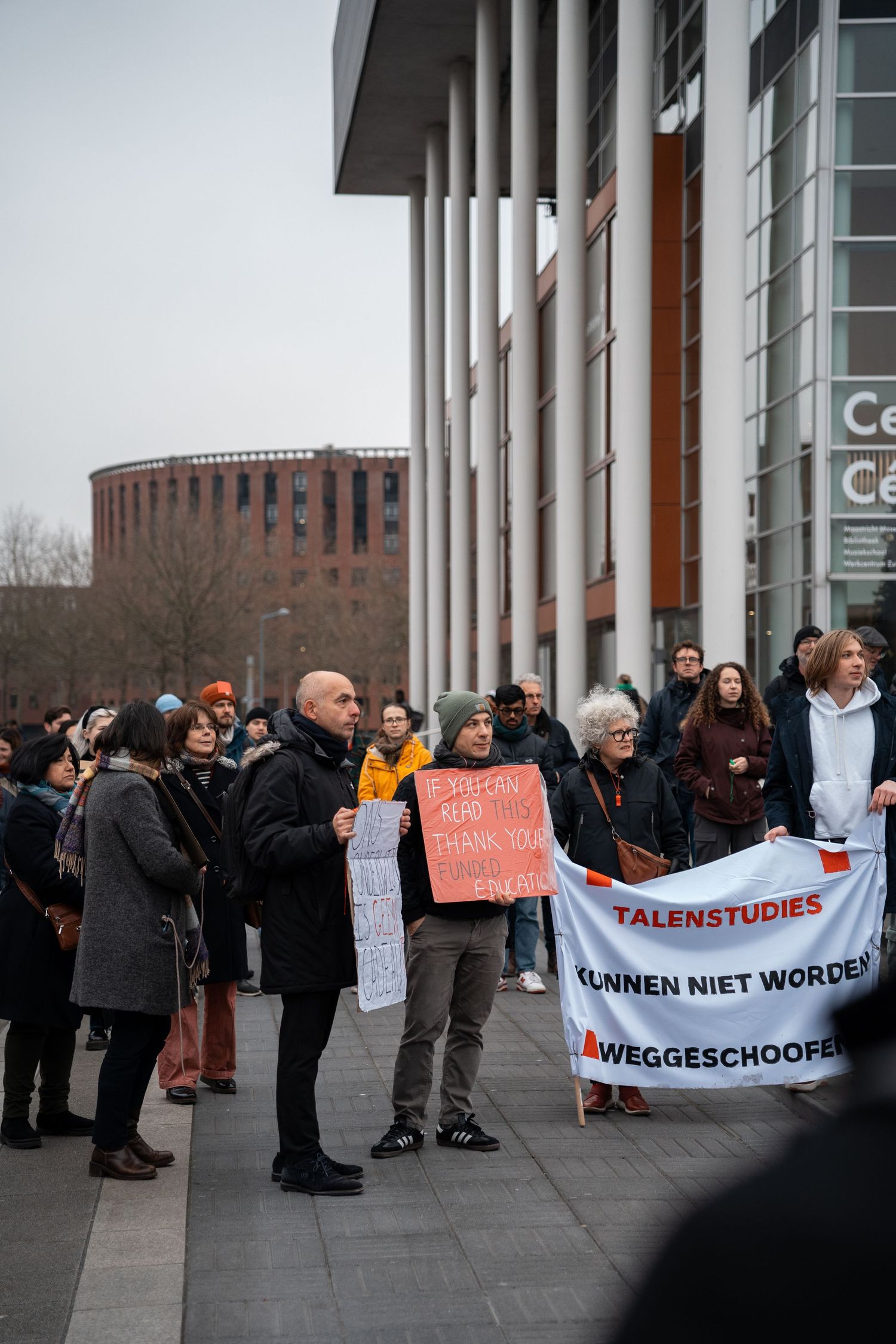 Protesters gather outside a building holding signs including a banner reading Talenstudies Kunnen Niet Worden Weggeschooid.