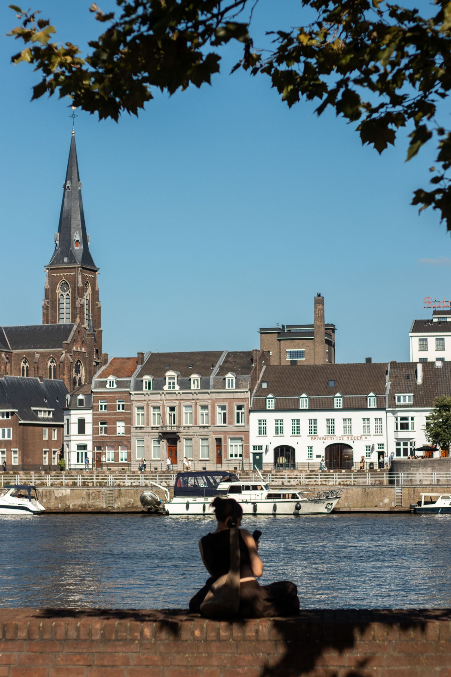 Person sitting on a riverside wall photographing a church spire and historic buildings across the Maas river.