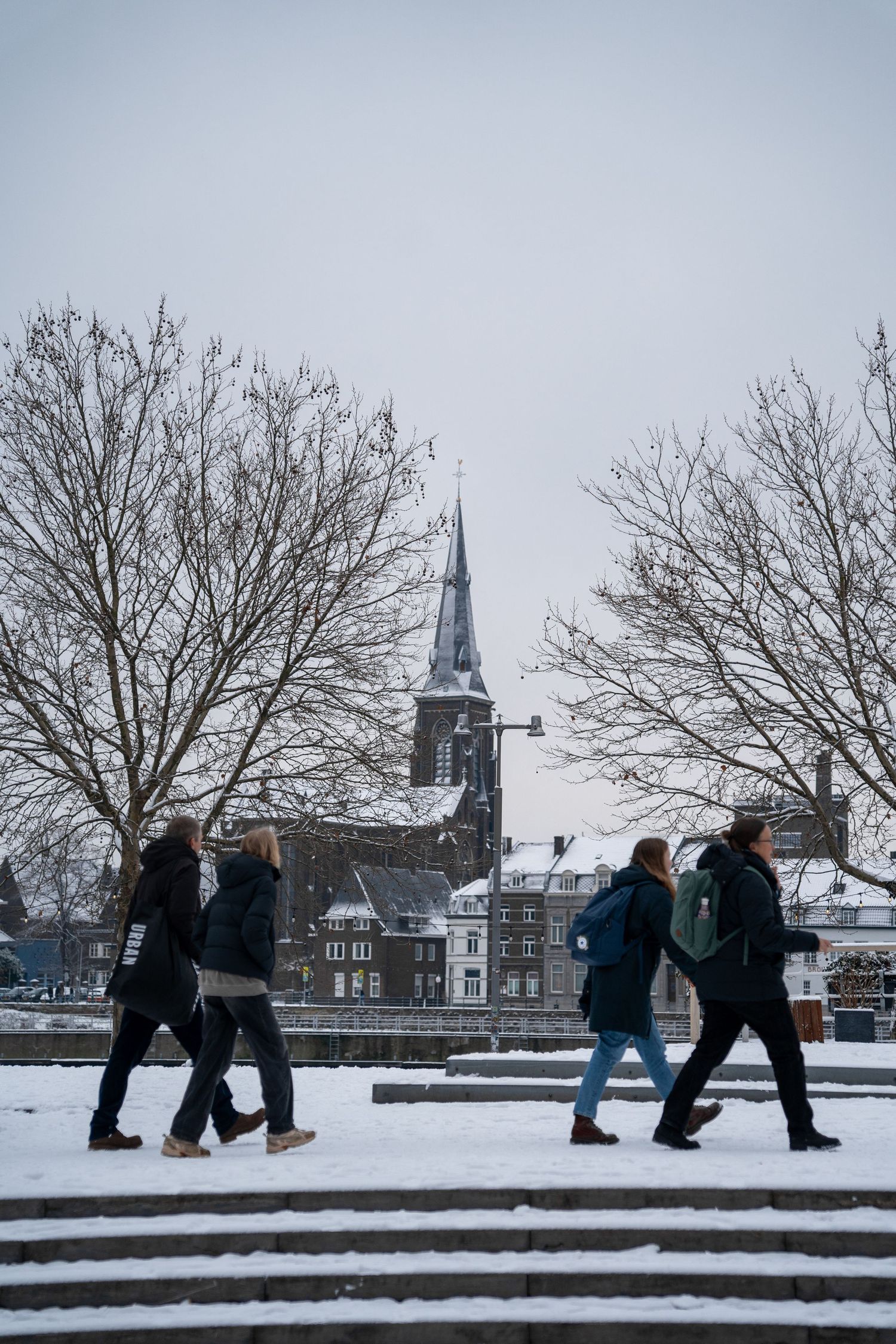People walking through snow past bare trees with a historic church steeple visible in a European city.