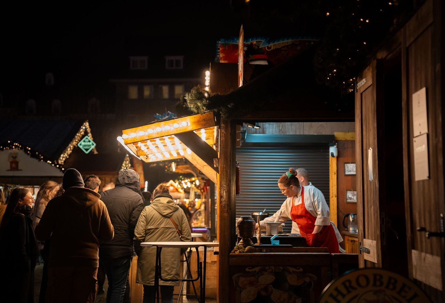 Vendor serving food at a festive Christmas market stall at night, with warm lights illuminating the wooden booth.