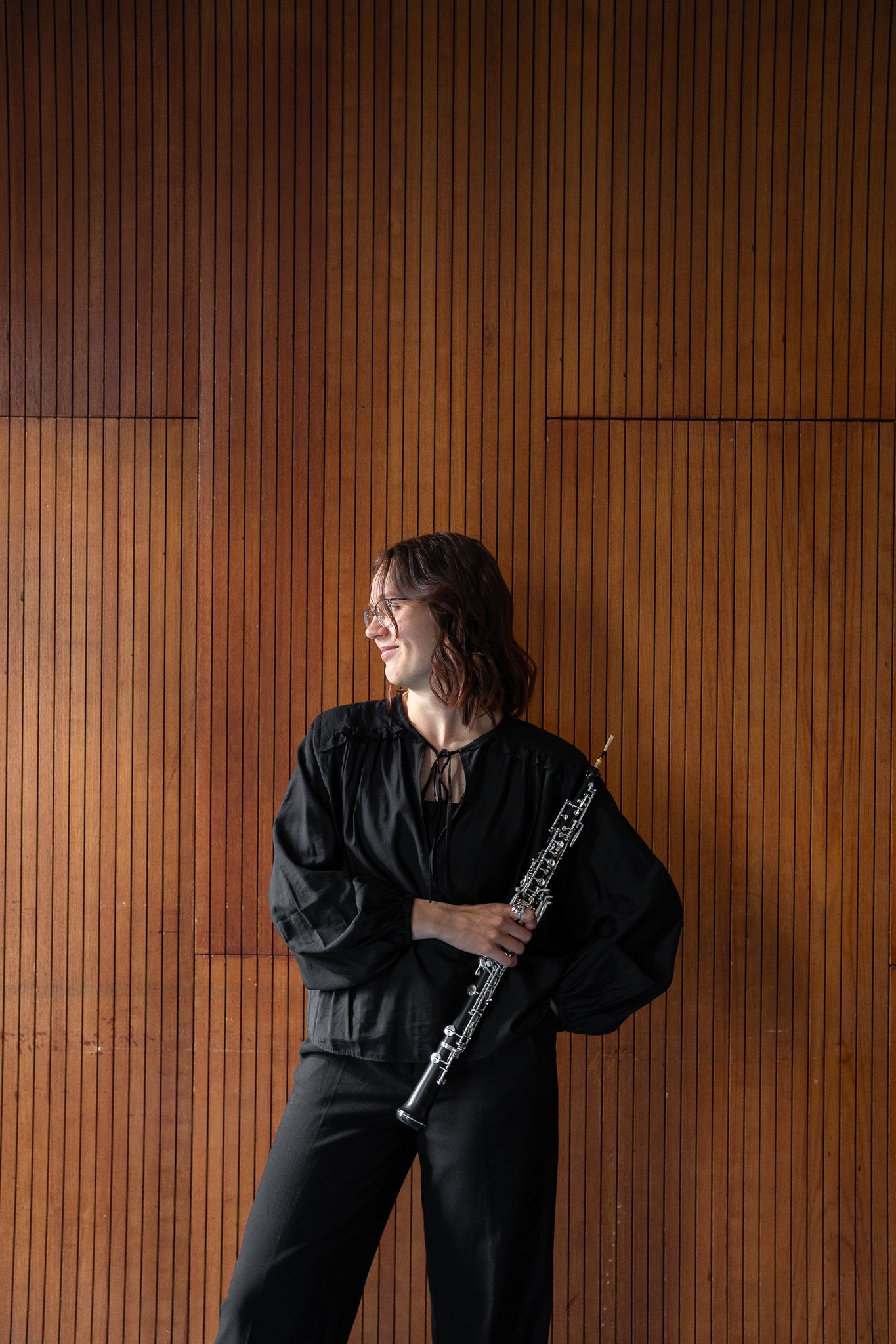 Female musician in black outfit holding an oboe while standing against a wooden panel wall.