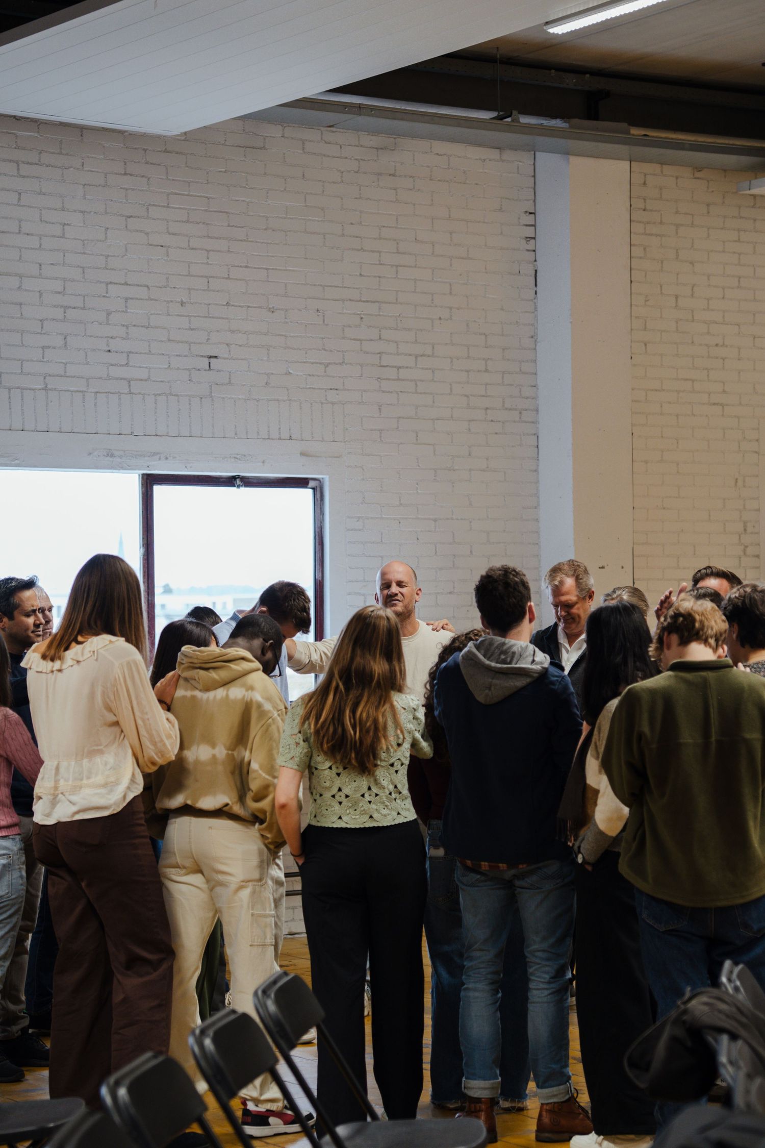 Group of people gathered in circle for prayer in modern white brick industrial church space.