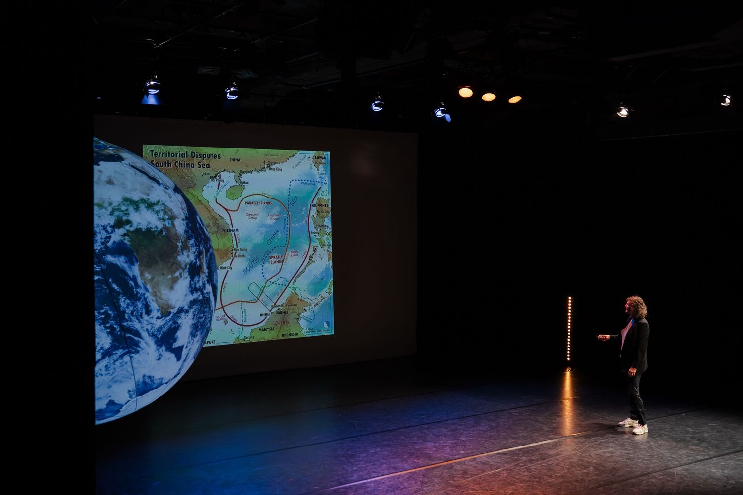 Performer on dark stage beside large projected globe and map display in black box theater.