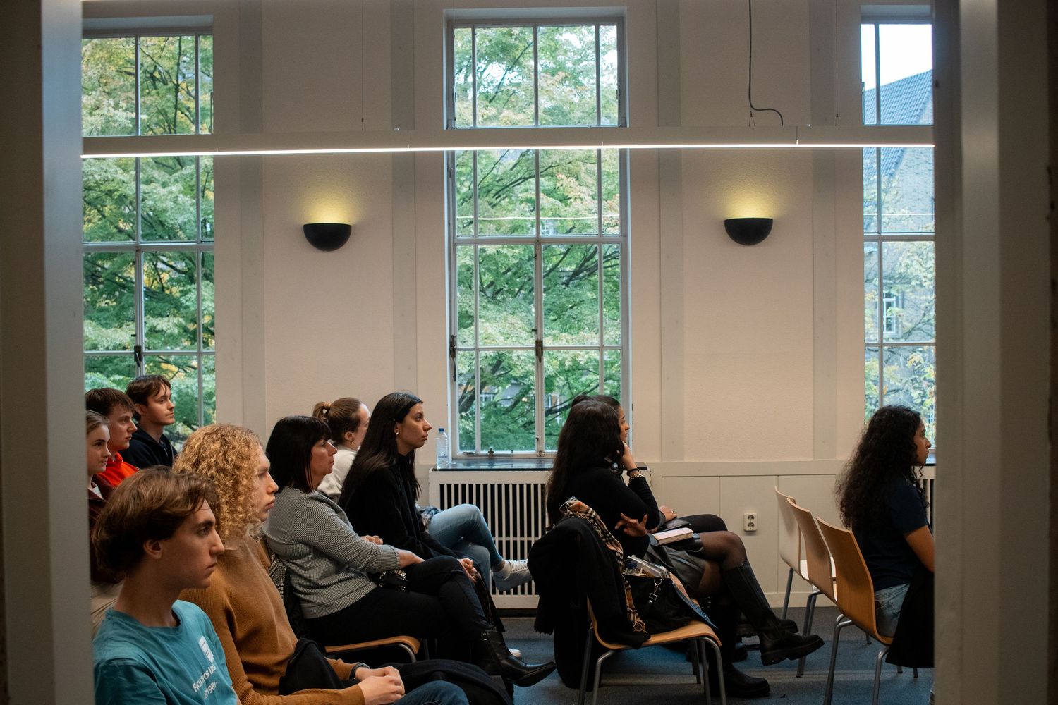 Students sit in rows of chairs during a university presentation in a bright room with tall windows.
