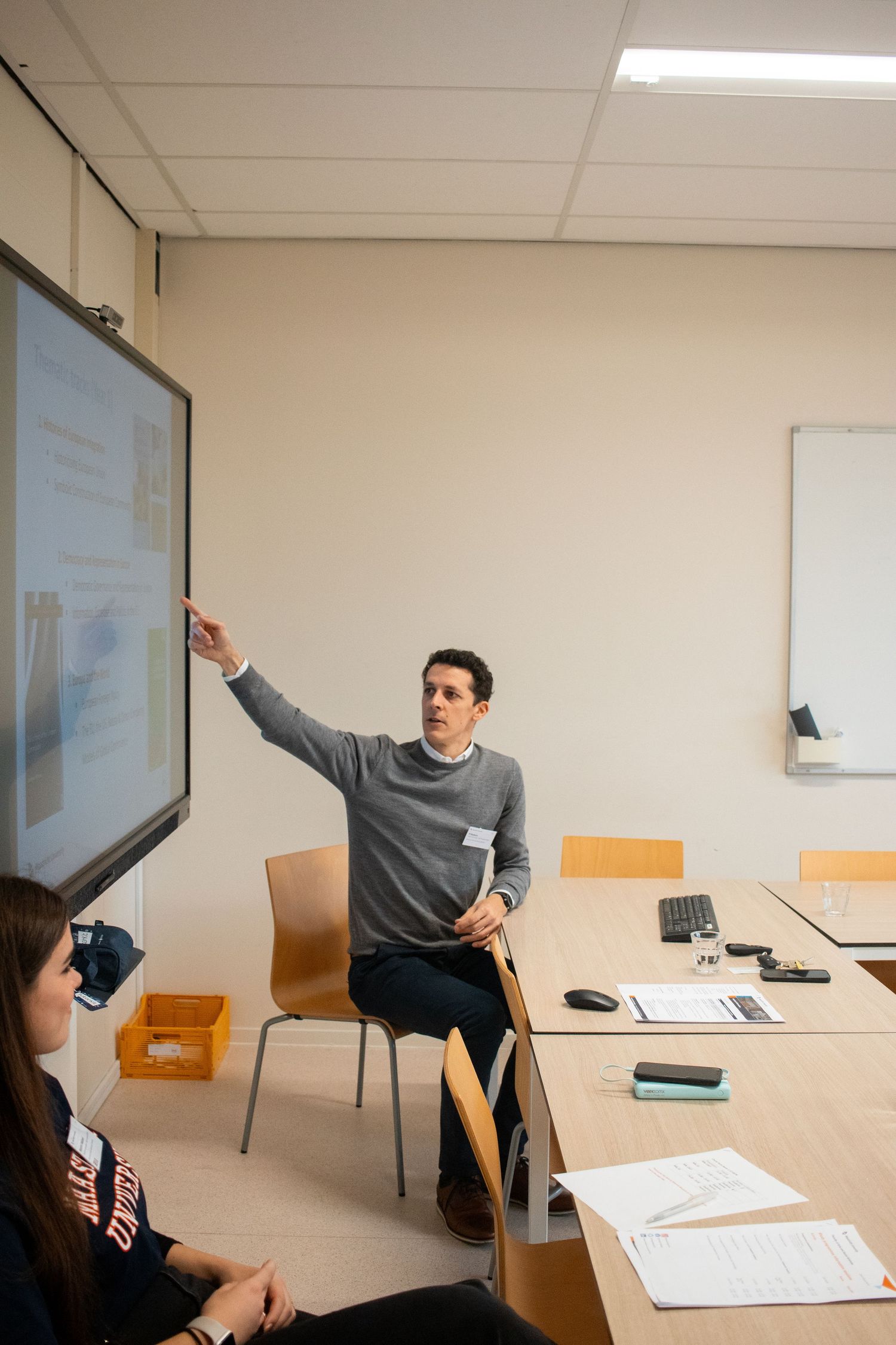 Man presenting at interactive whiteboard in modern classroom with student audience.