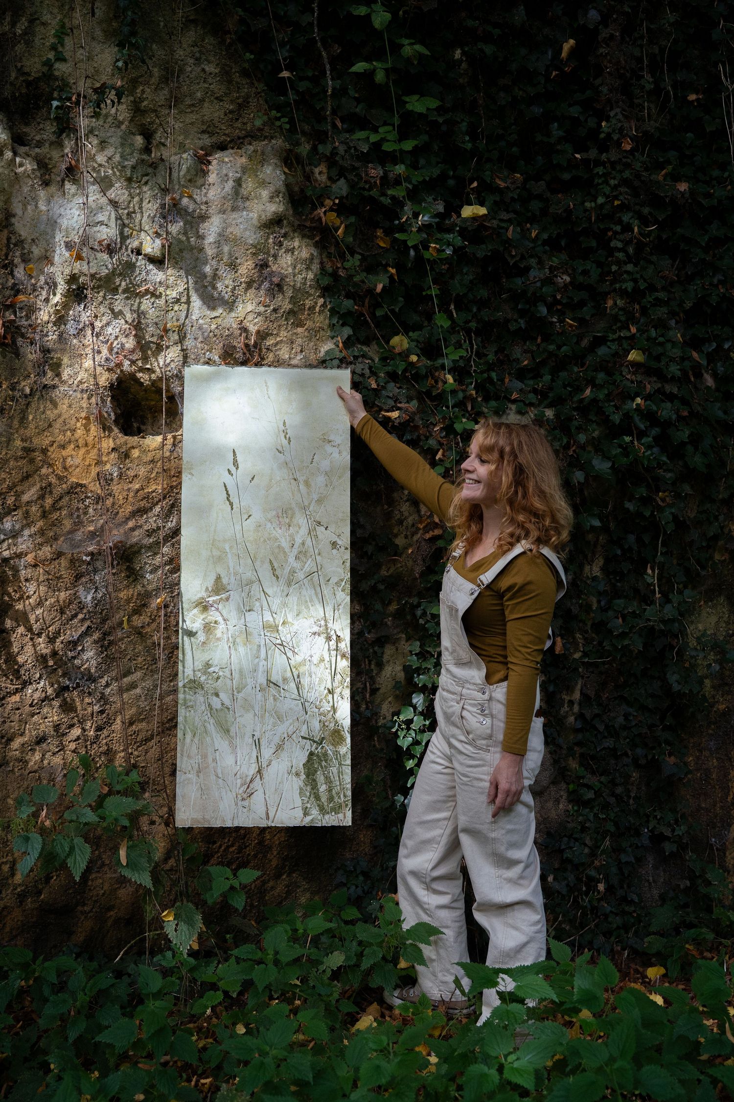 Woman in overalls smiling while holding a rectangular botanical art panel against a mossy rock wall.