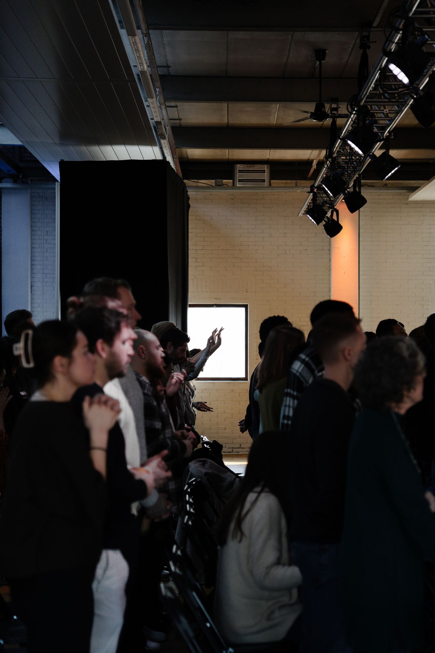 Congregation members standing and worshipping in a dimly lit modern church venue with stage lighting.
