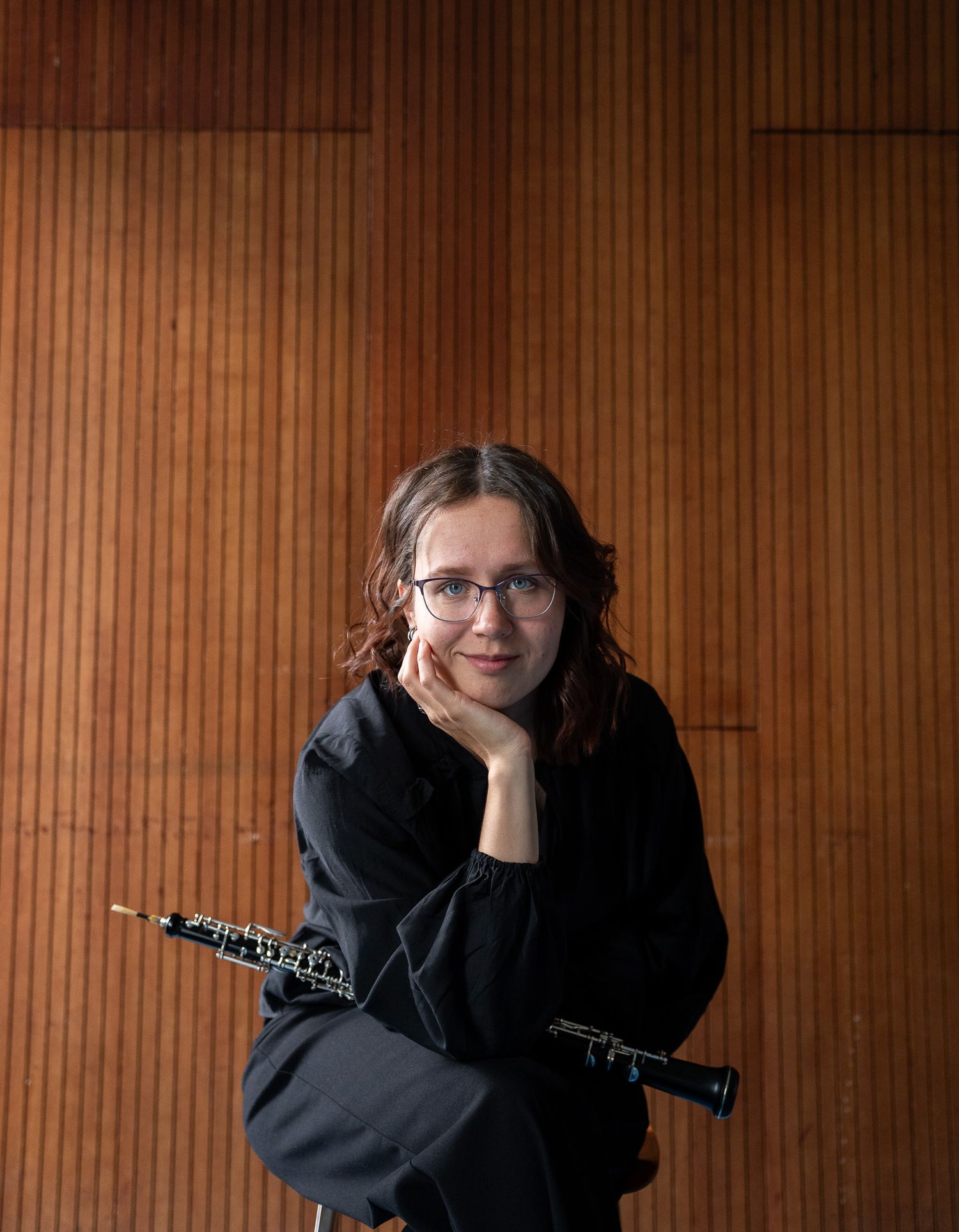 Female oboist in black outfit leaning against a wooden wall holding her oboe with chin resting on hand.