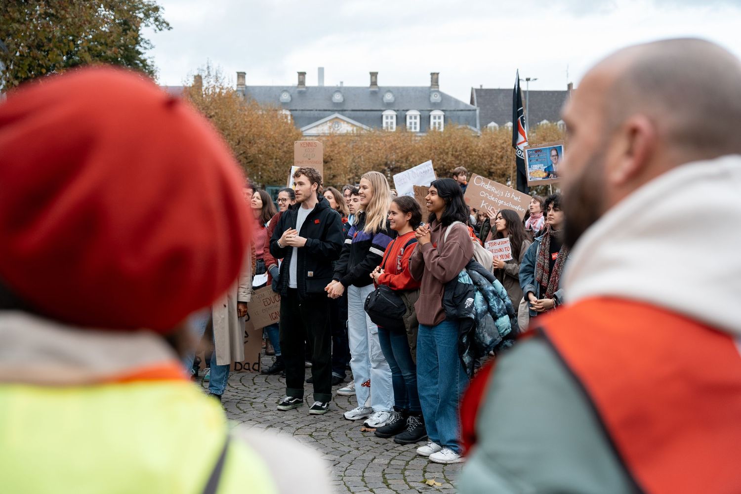 Students and protesters gather attentively at outdoor rally with signs on autumn day.