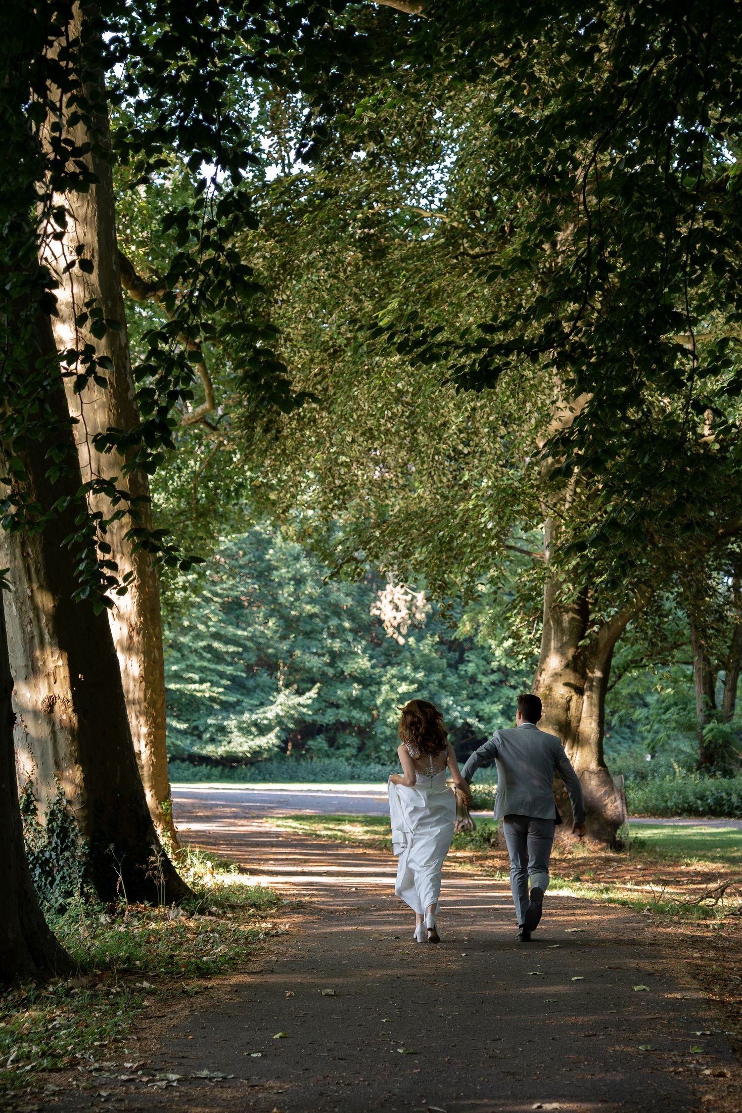 Bride and groom walking hand in hand down a sunlit tree-lined park path on their wedding day.