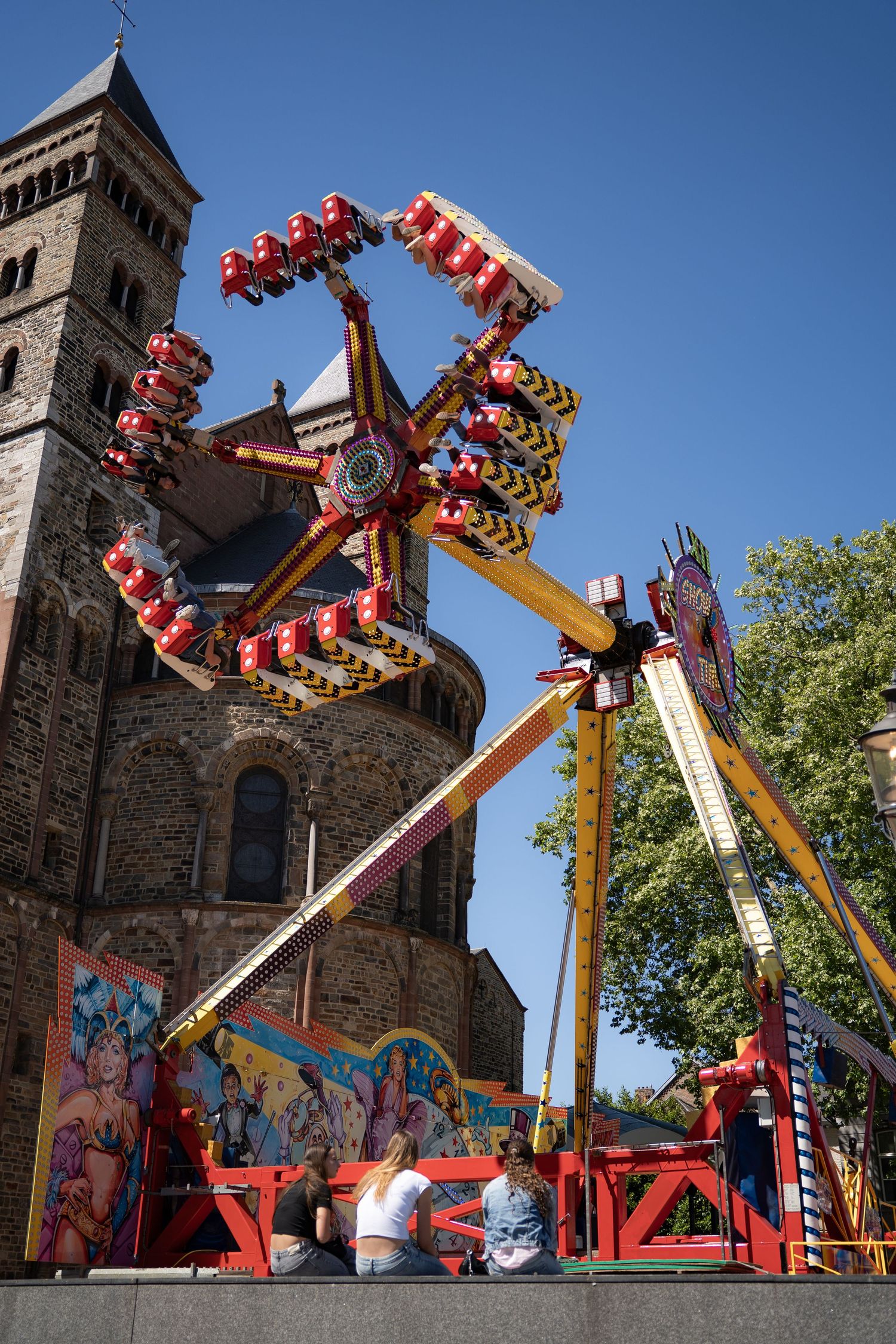 Colorful thrill ride spinning at a fairground with a historic stone church tower visible against a clear blue sky.