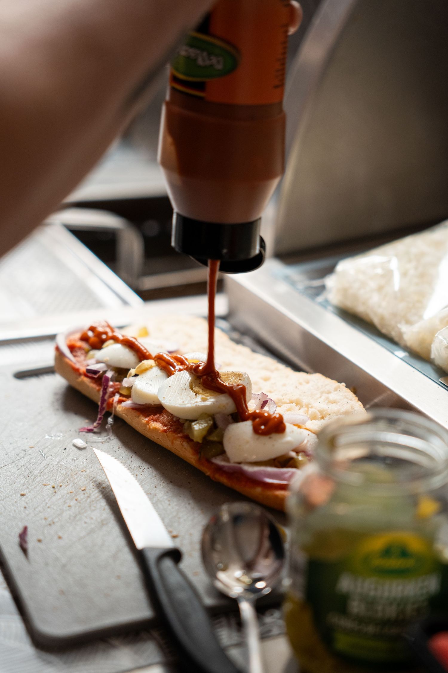 Ketchup being squeezed from a bottle onto an open-faced sandwich with cheese and toppings on a wooden cutting board.