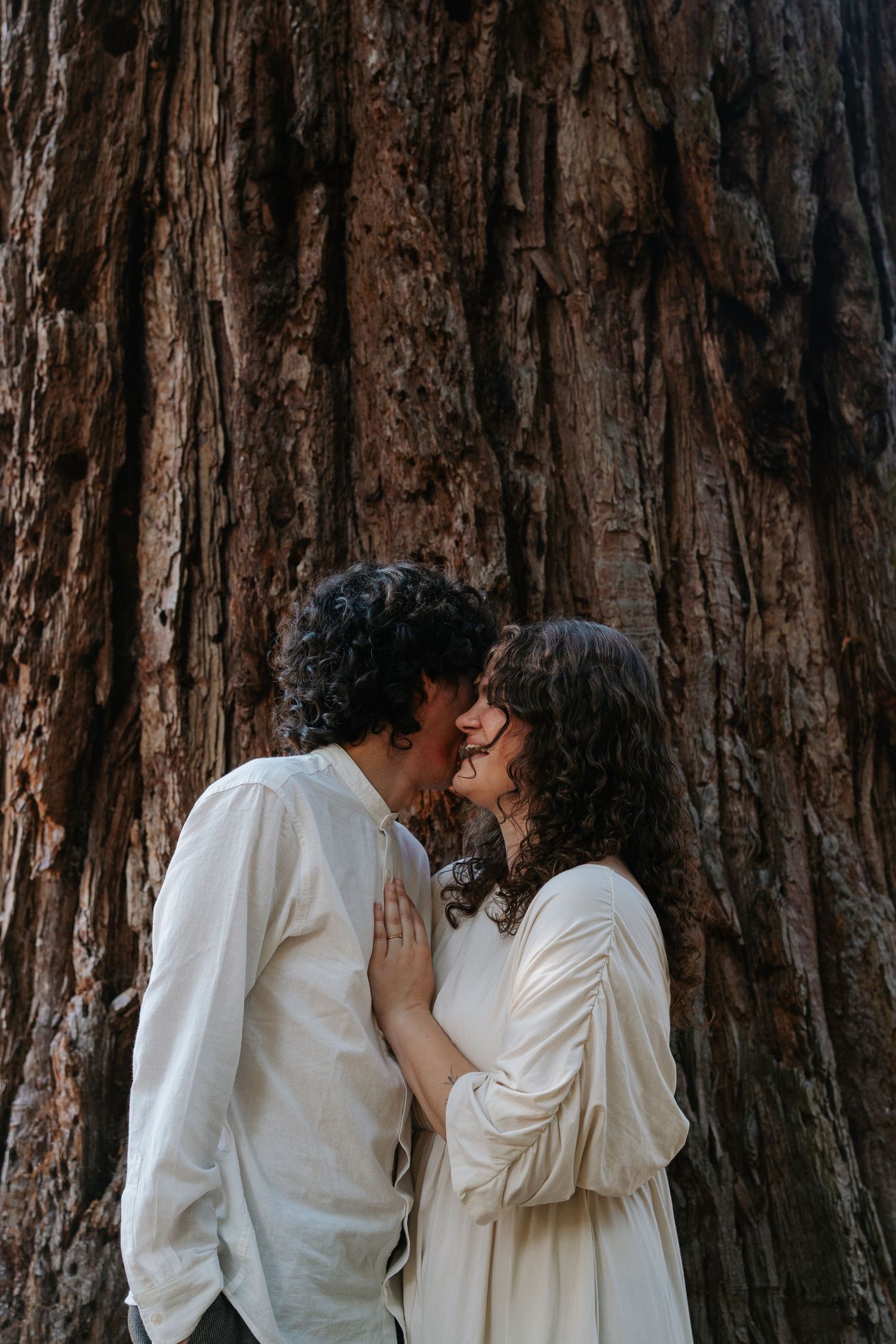 A couple in white clothing shares a tender kiss in front of a large redwood tree trunk.