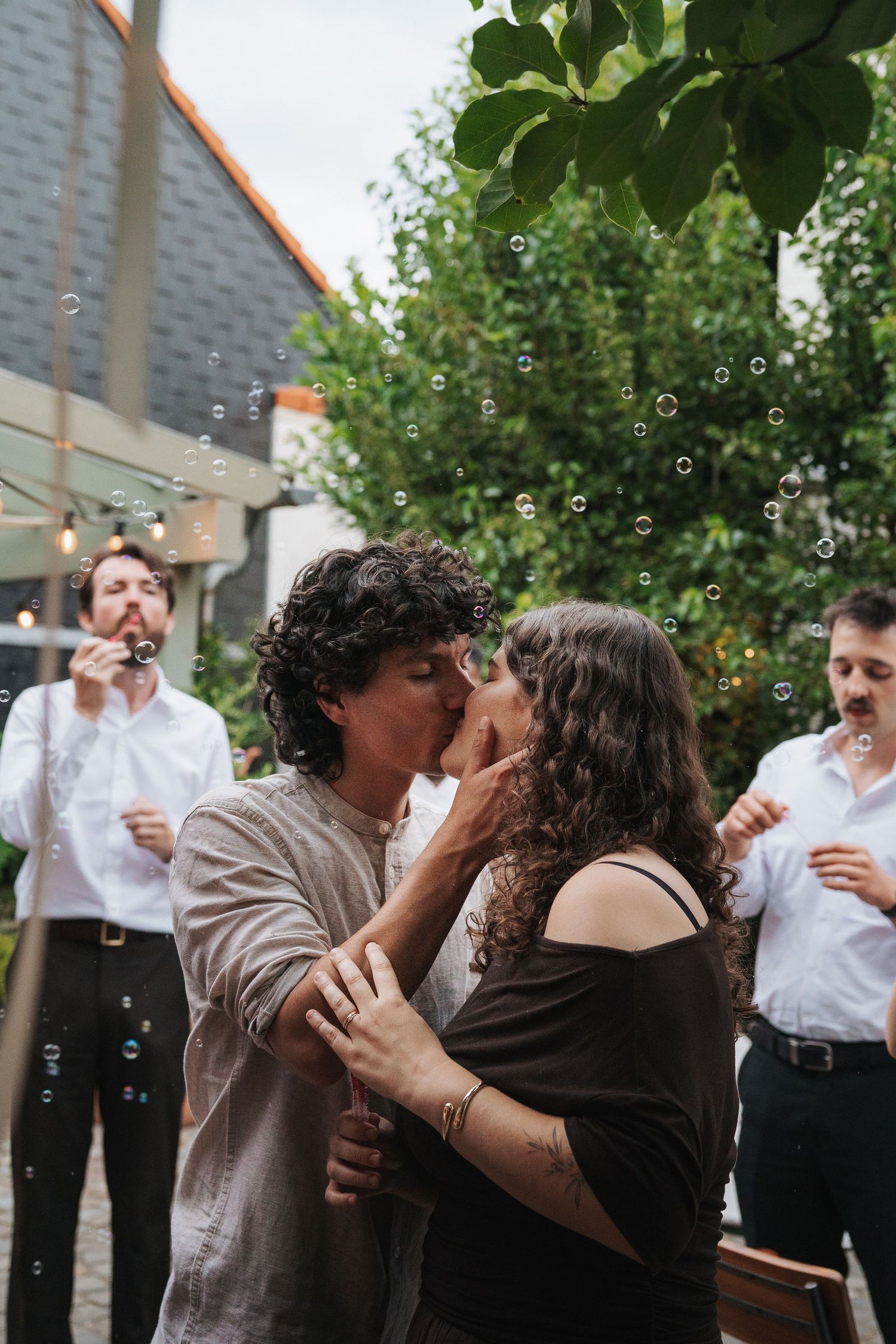 Couple sharing a romantic kiss surrounded by soap bubbles at an outdoor celebration with guests blowing bubbles behind them.