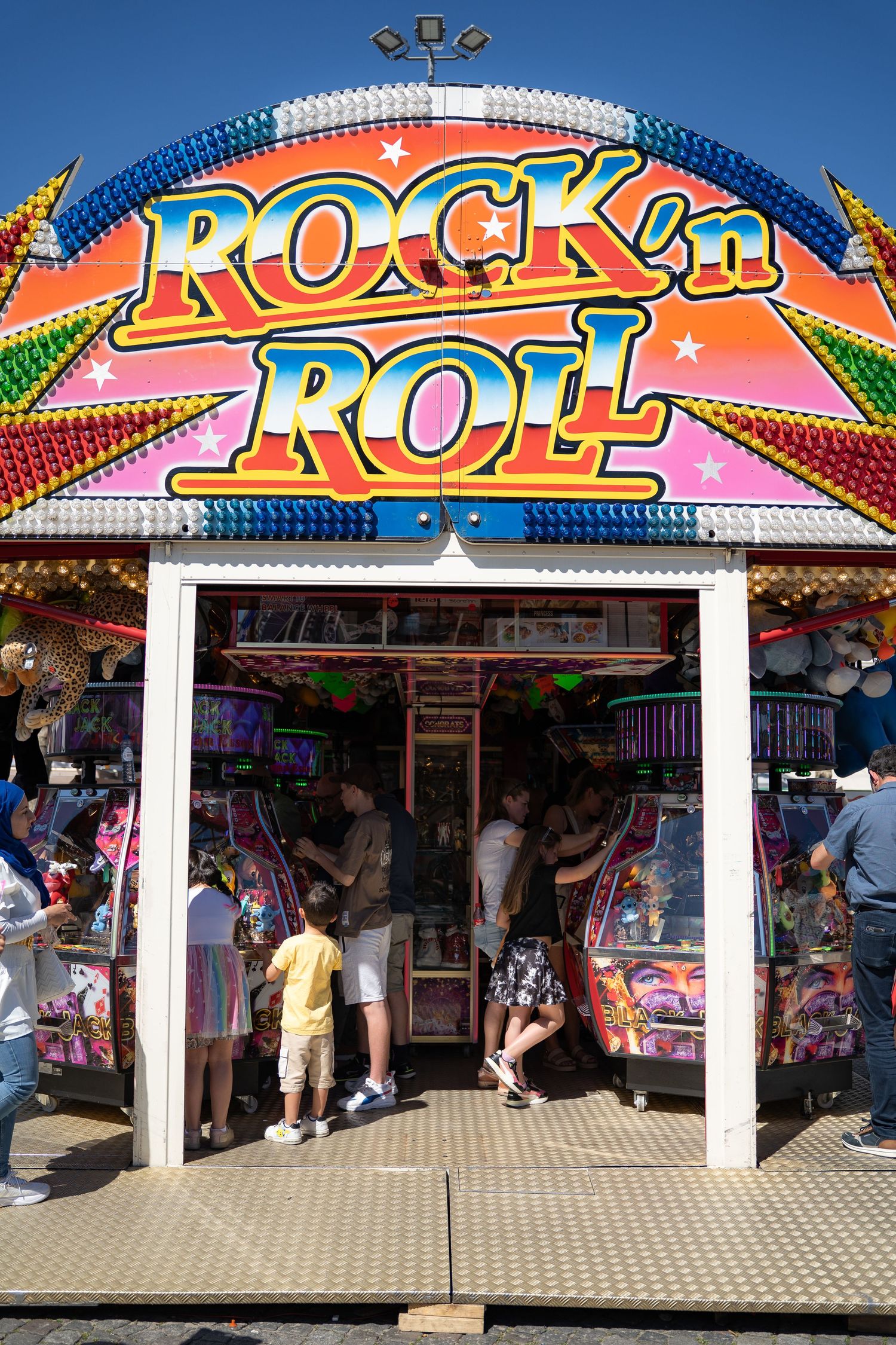 People enter a brightly lit Rock n Roll arcade game booth at a lively outdoor fairground on a sunny day.
