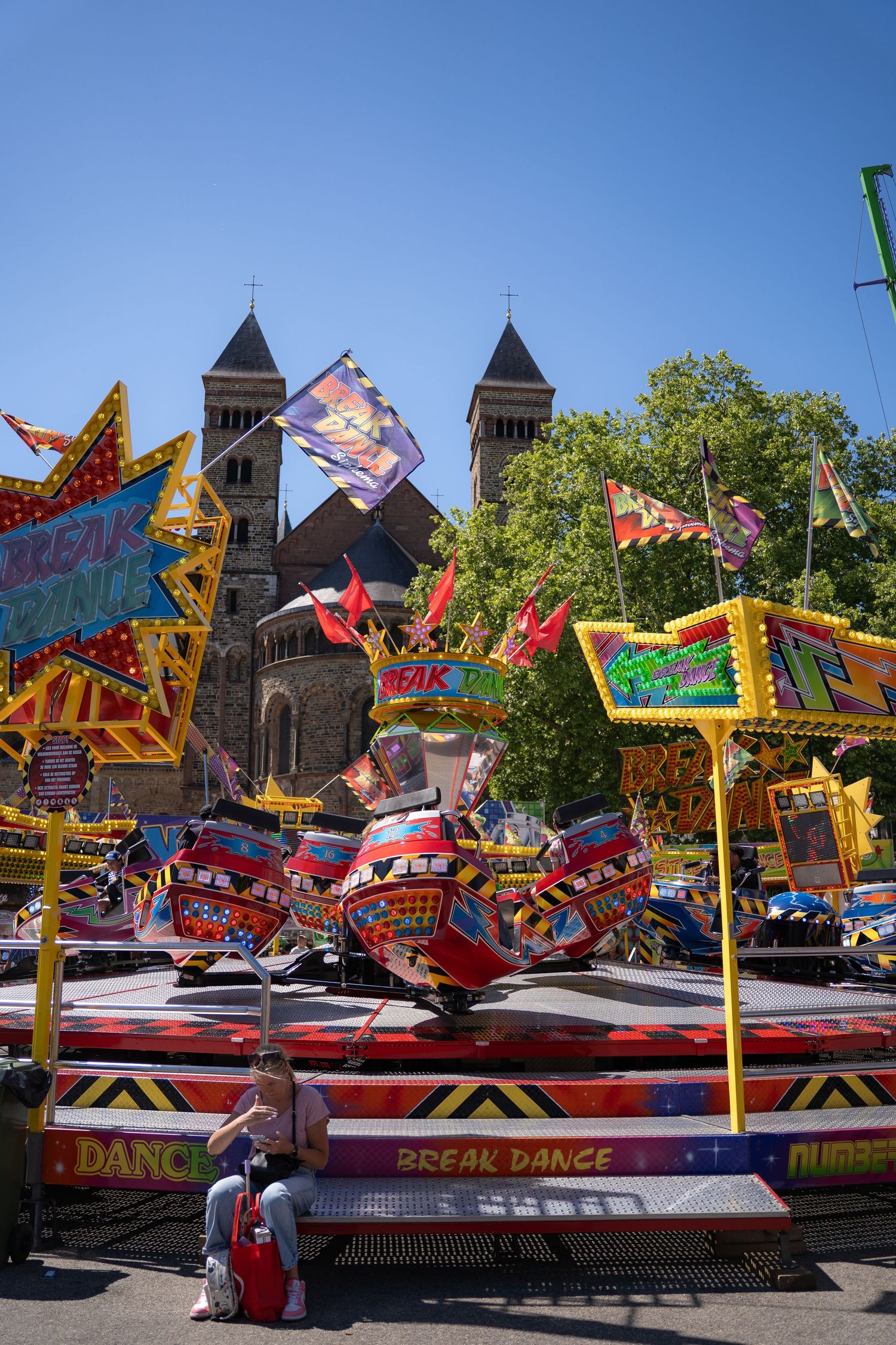 A colorful Break Dance carnival ride with flags and spinning cars near a historic twin-towered church on a sunny day.