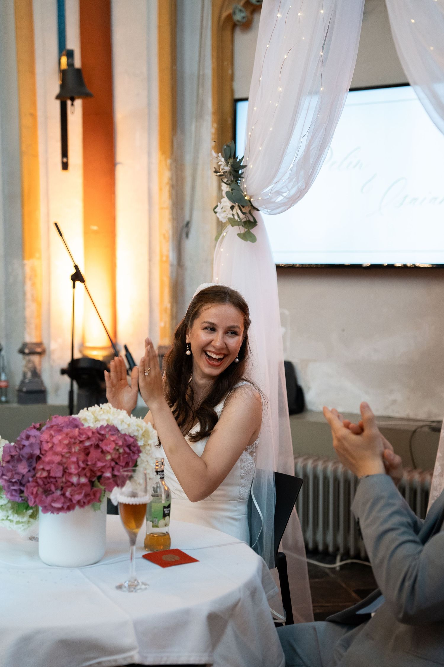 Smiling bride in white dress claps at the wedding reception table adorned with purple hydrangeas.