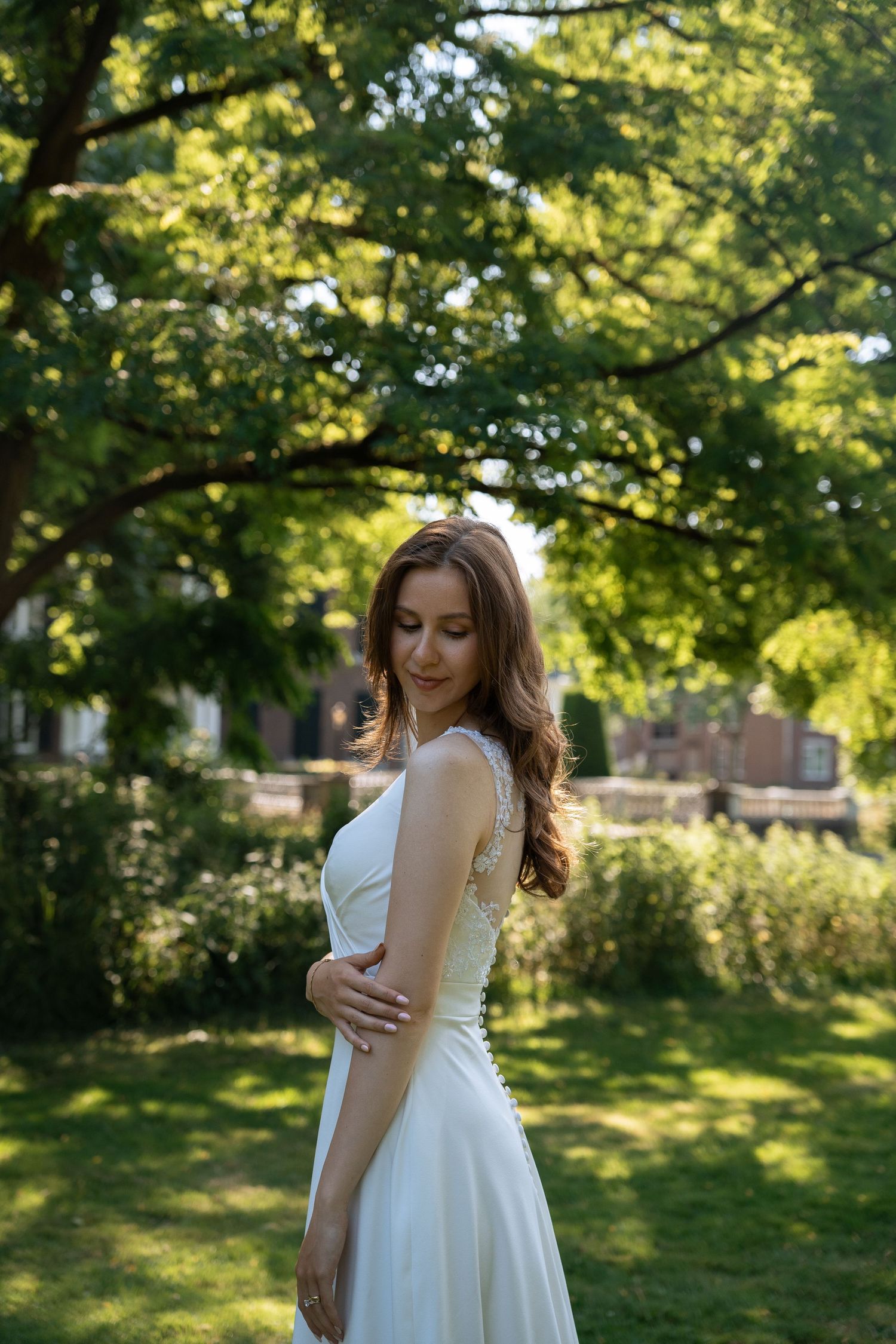 Elegant bride in white gown standing in a sunlit garden with lush green trees.