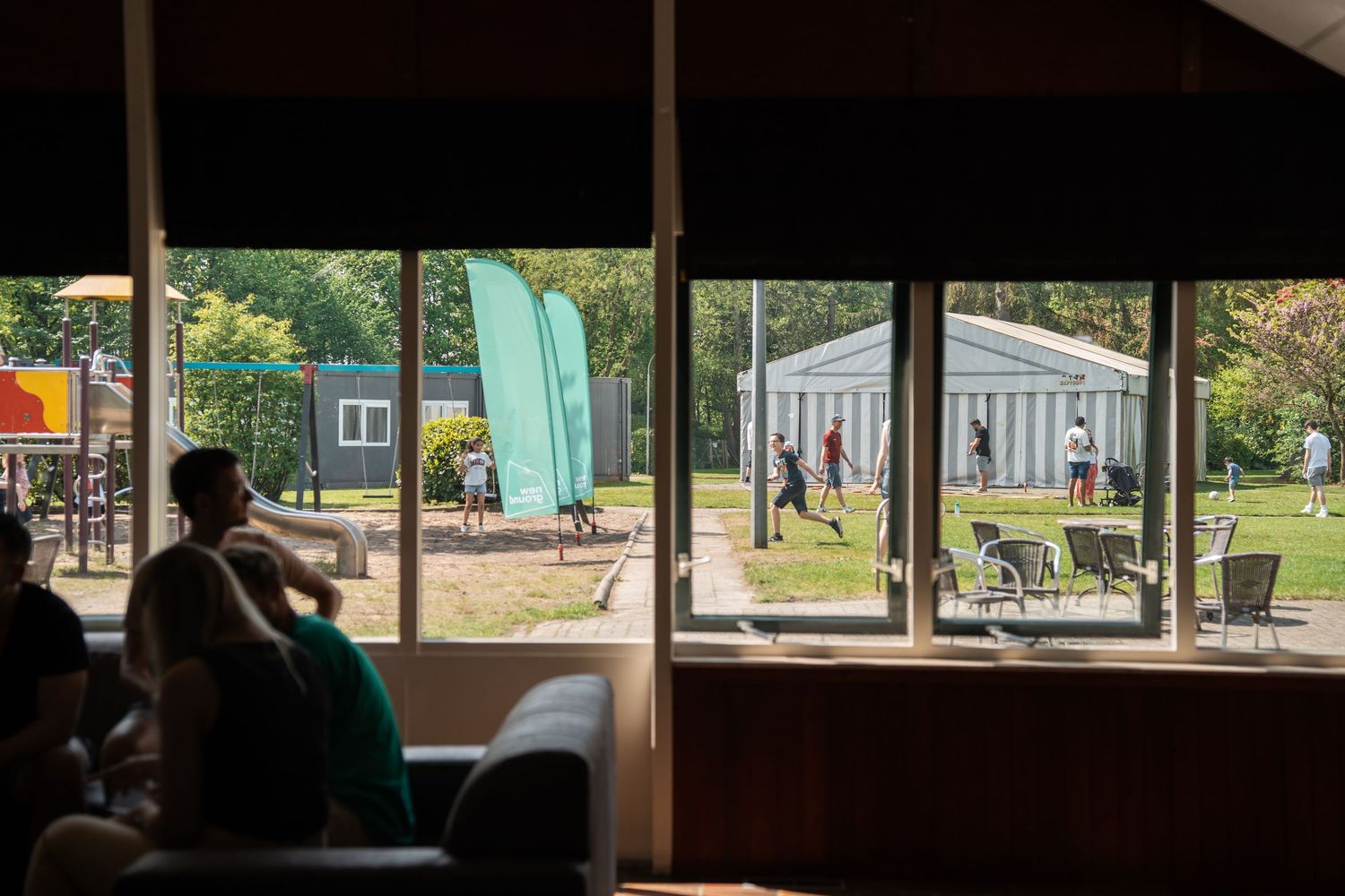 View through large windows of an outdoor venue with green banner flags and people walking outside.