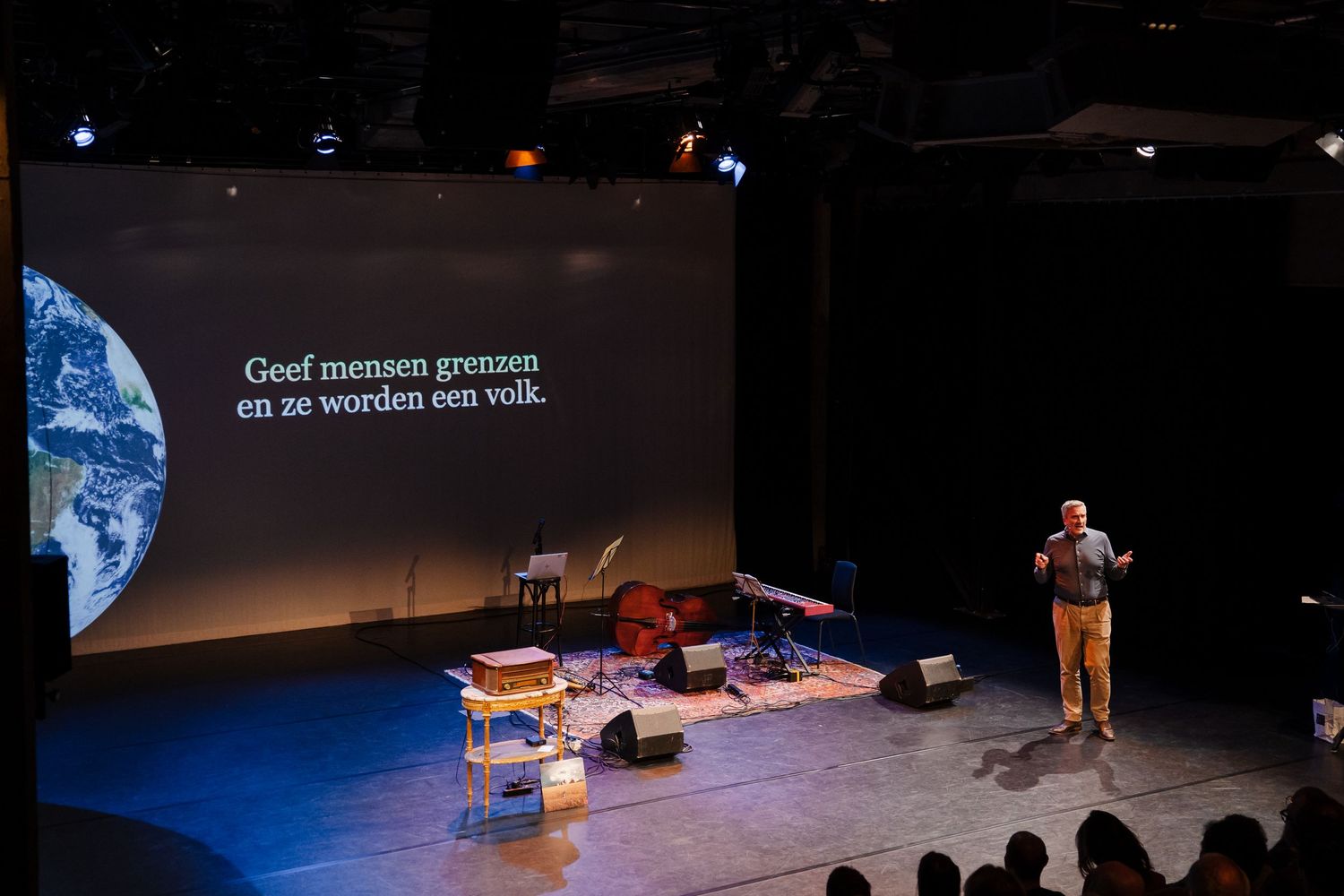 Speaker on stage with Dutch text projection and Earth image at academic event.
