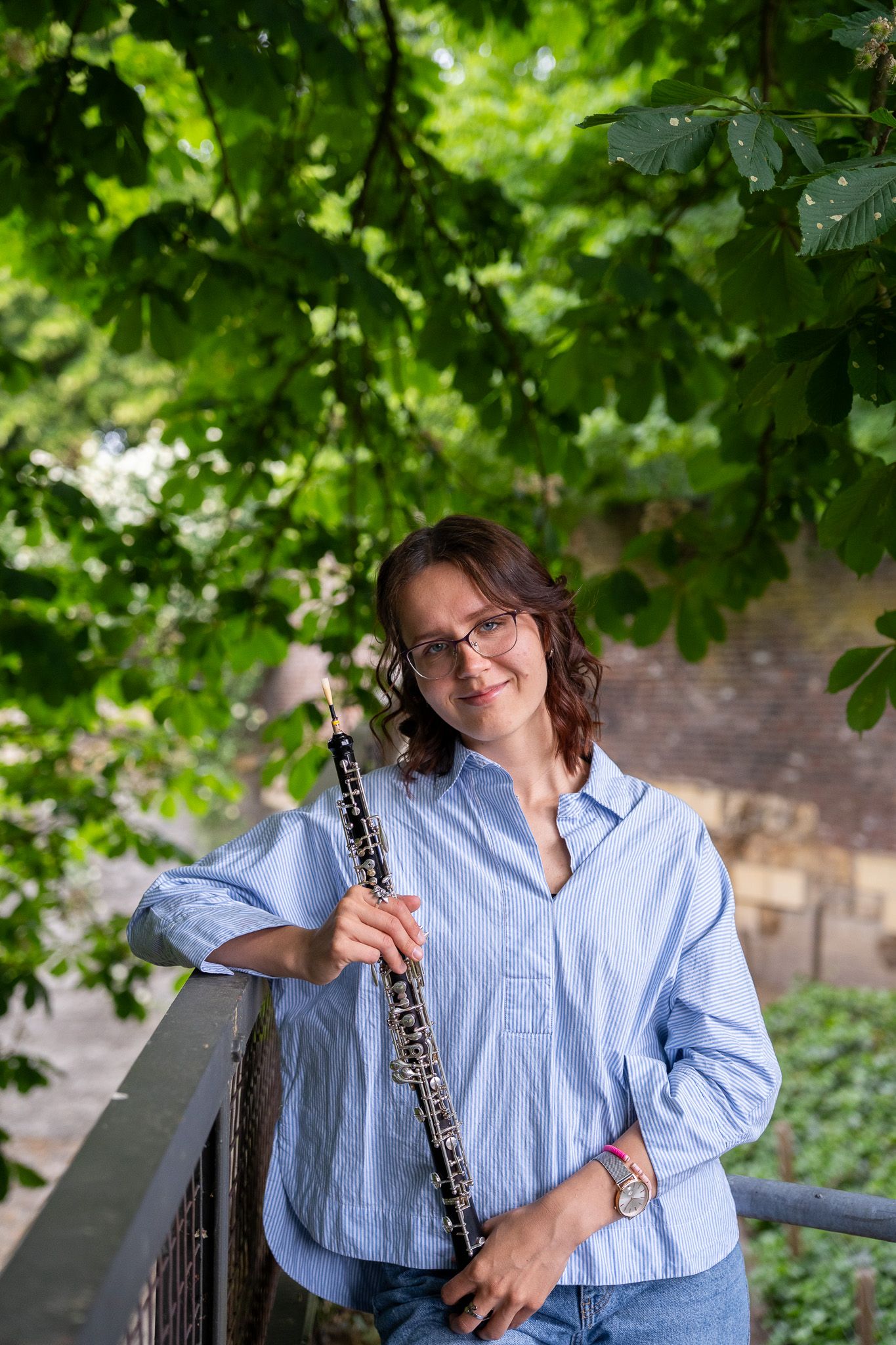 Smiling female oboist in a blue shirt holding her oboe outdoors under lush green tree branches.
