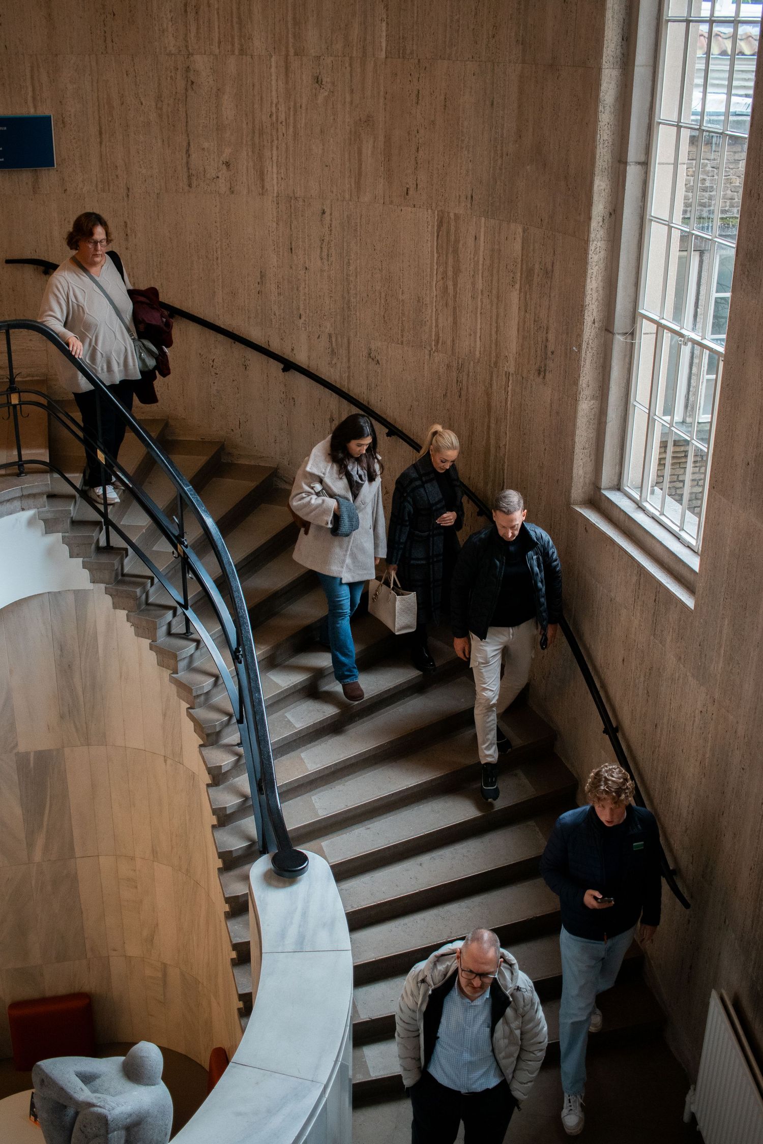 Visitors descending a curved marble staircase inside a modern museum with high stone walls and large windows.