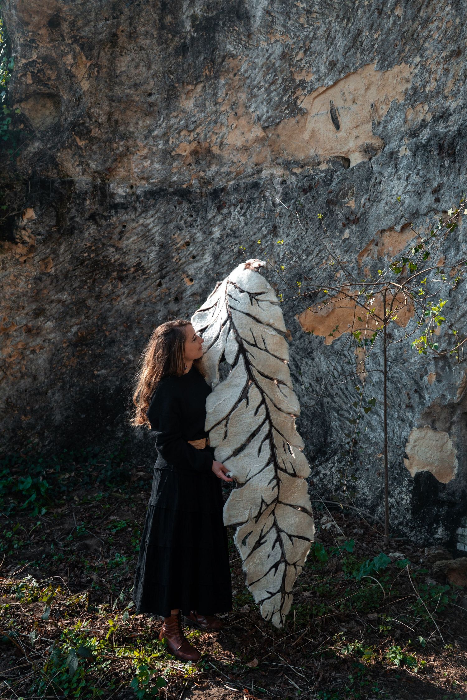Woman in black dress hugging a large white leaf sculpture against a weathered stone wall.