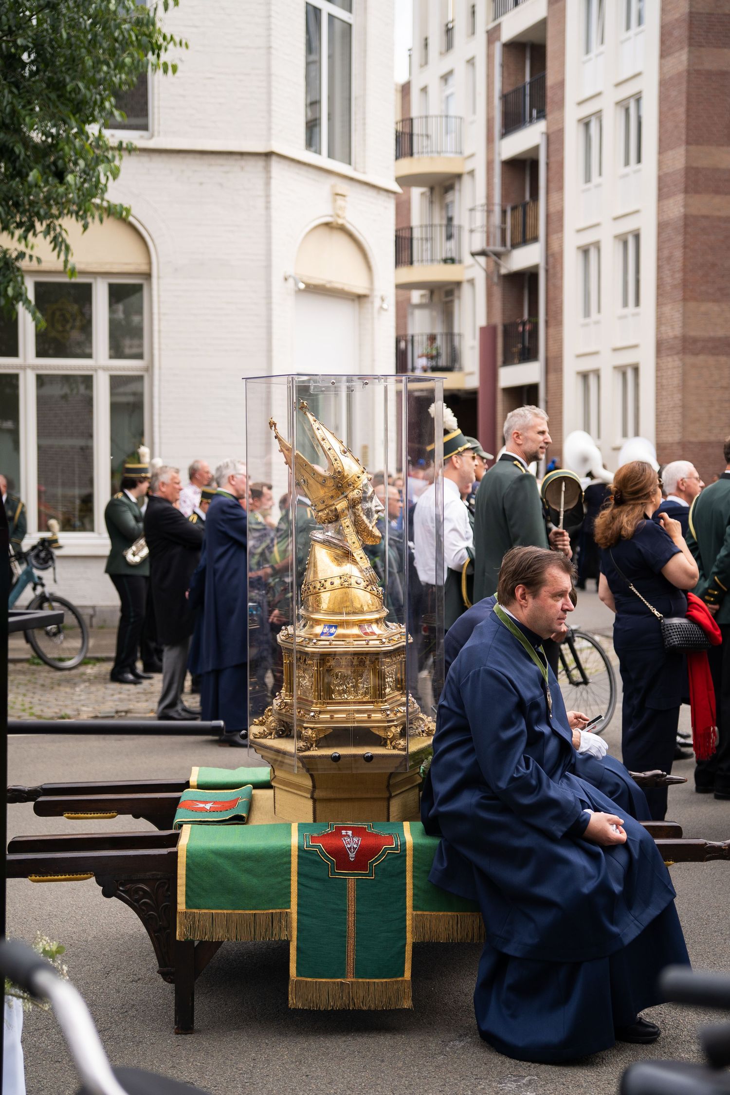 Man in blue robe sits beside an ornate golden religious reliquary on a cart during a procession.