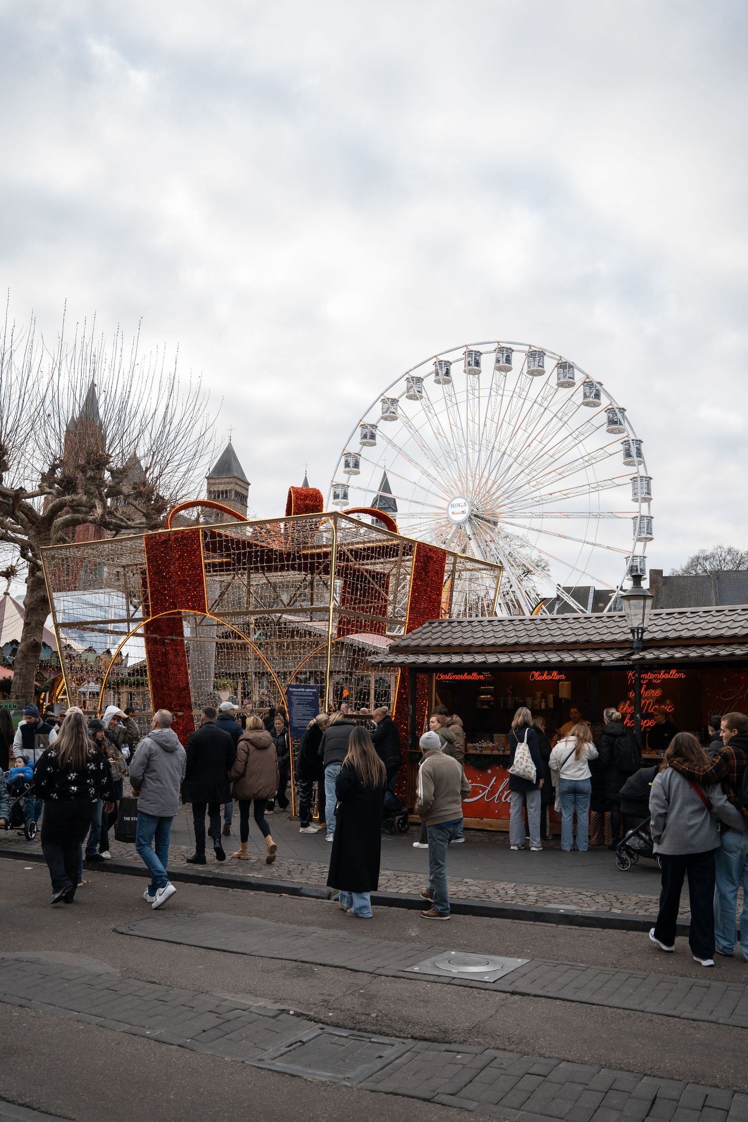 Crowds exploring a festive Christmas market with a large Ferris wheel and red gift box decoration.