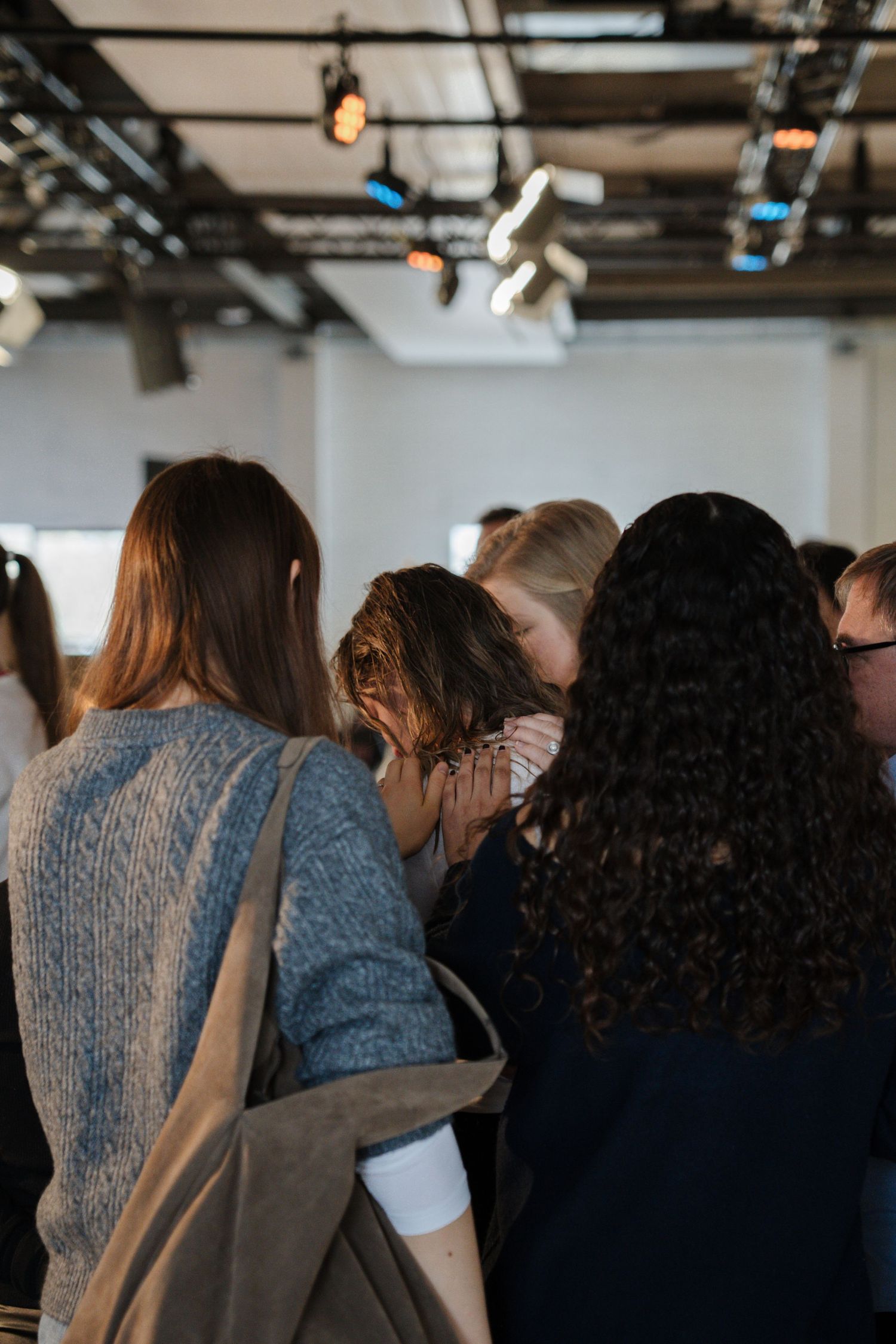 Group of women standing close together in prayer at an indoor religious gathering with stage lighting above.