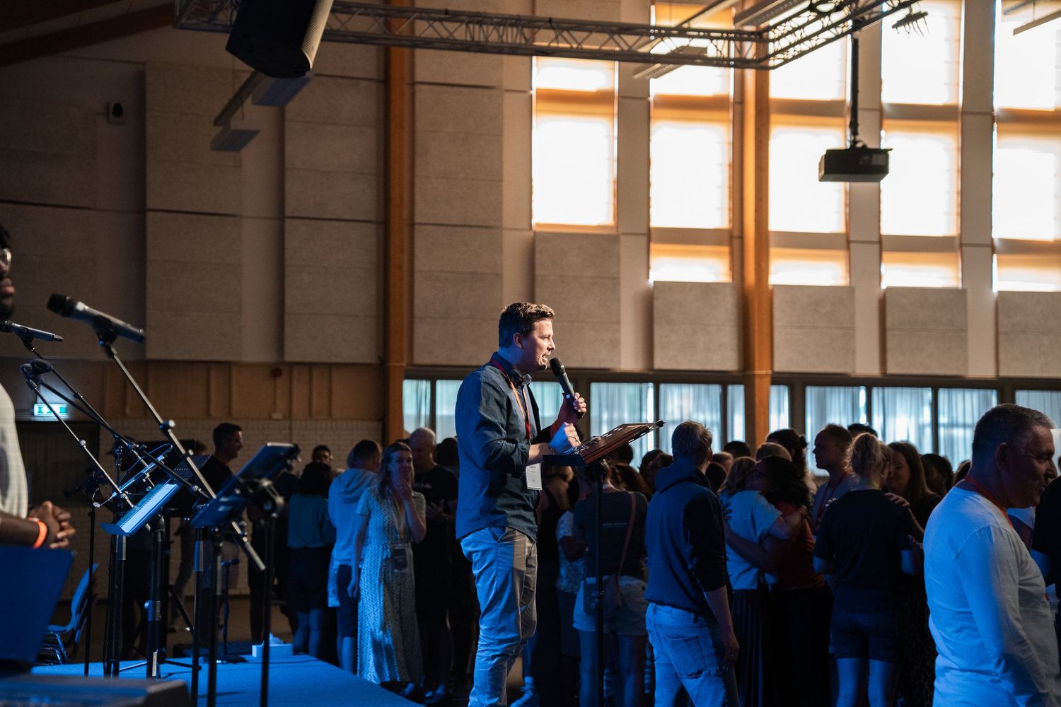 Speaker addresses a standing congregation holding a microphone during a sunlit Christian worship service.