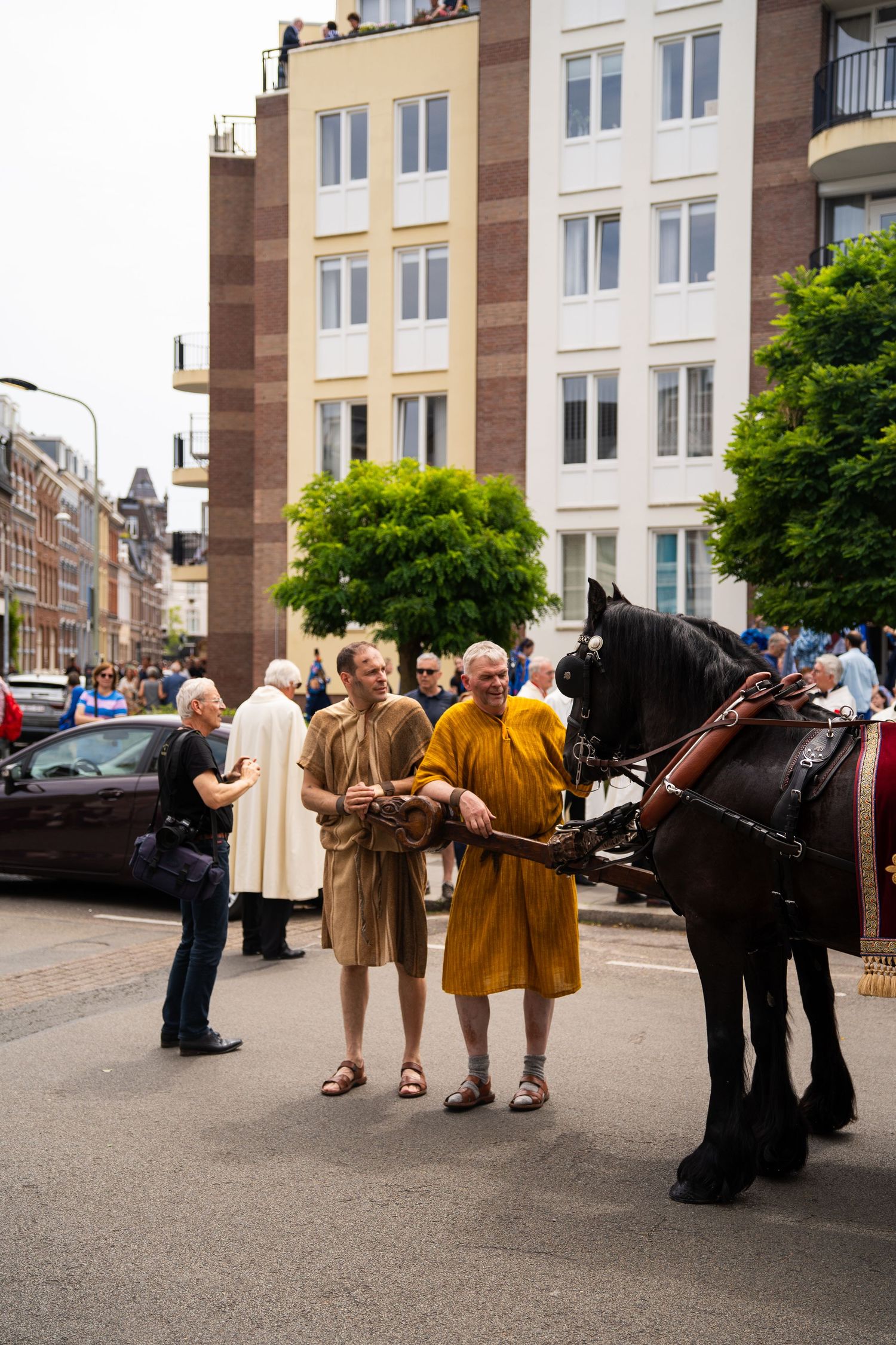 Two men dressed in historical costumes stand beside a black horse during a street parade.
