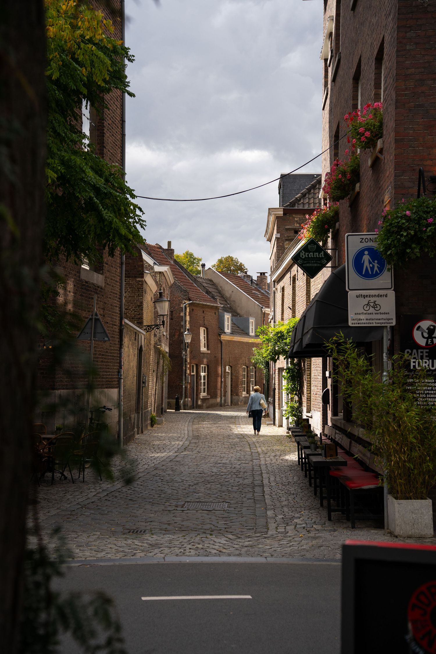 Narrow cobblestone street lined with brick buildings, flower boxes, and a cafe in a historic European town.