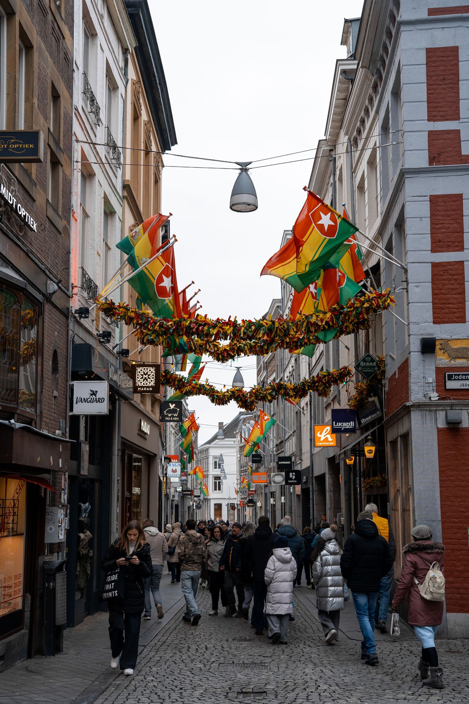 Shoppers walk through a festive European cobblestone street decorated with colorful flags and garland overhead.