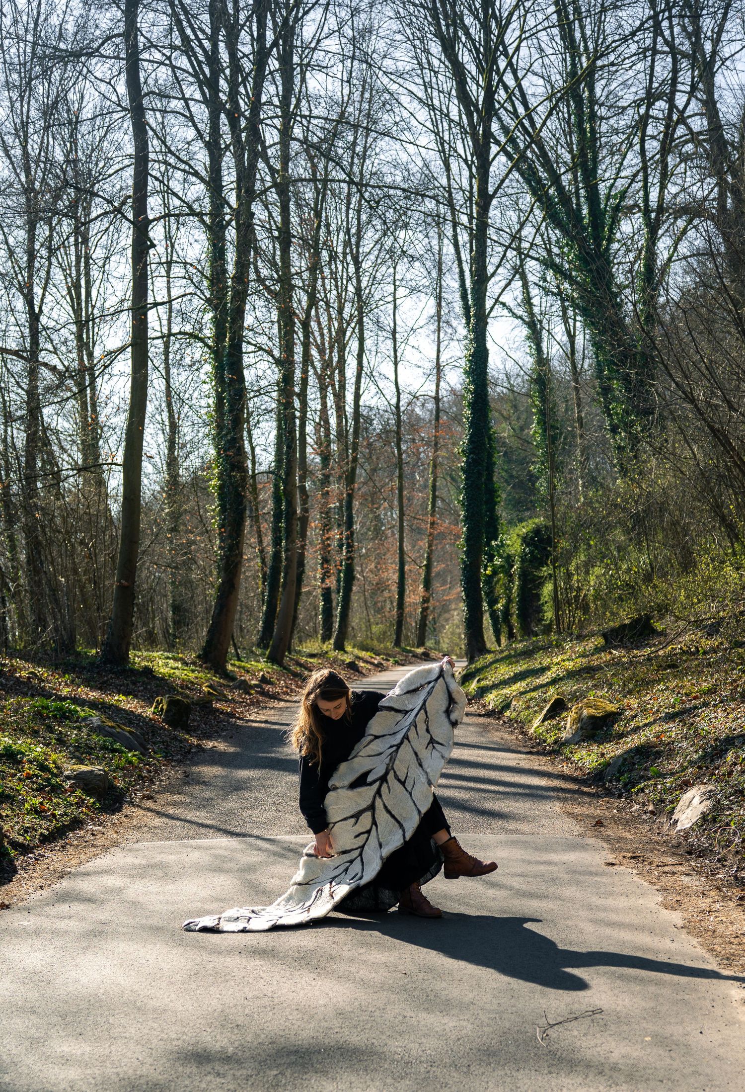 Woman in black holding a large white leaf-patterned textile on a sunlit forest road.