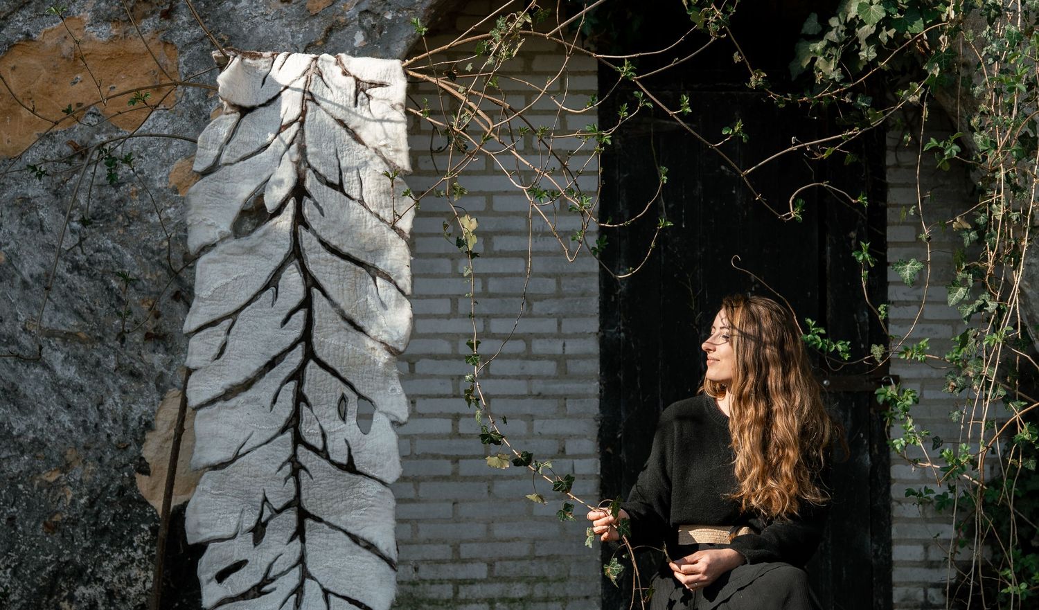 Woman in black outfit standing beside a tall white leaf sculpture against a brick wall outdoors.