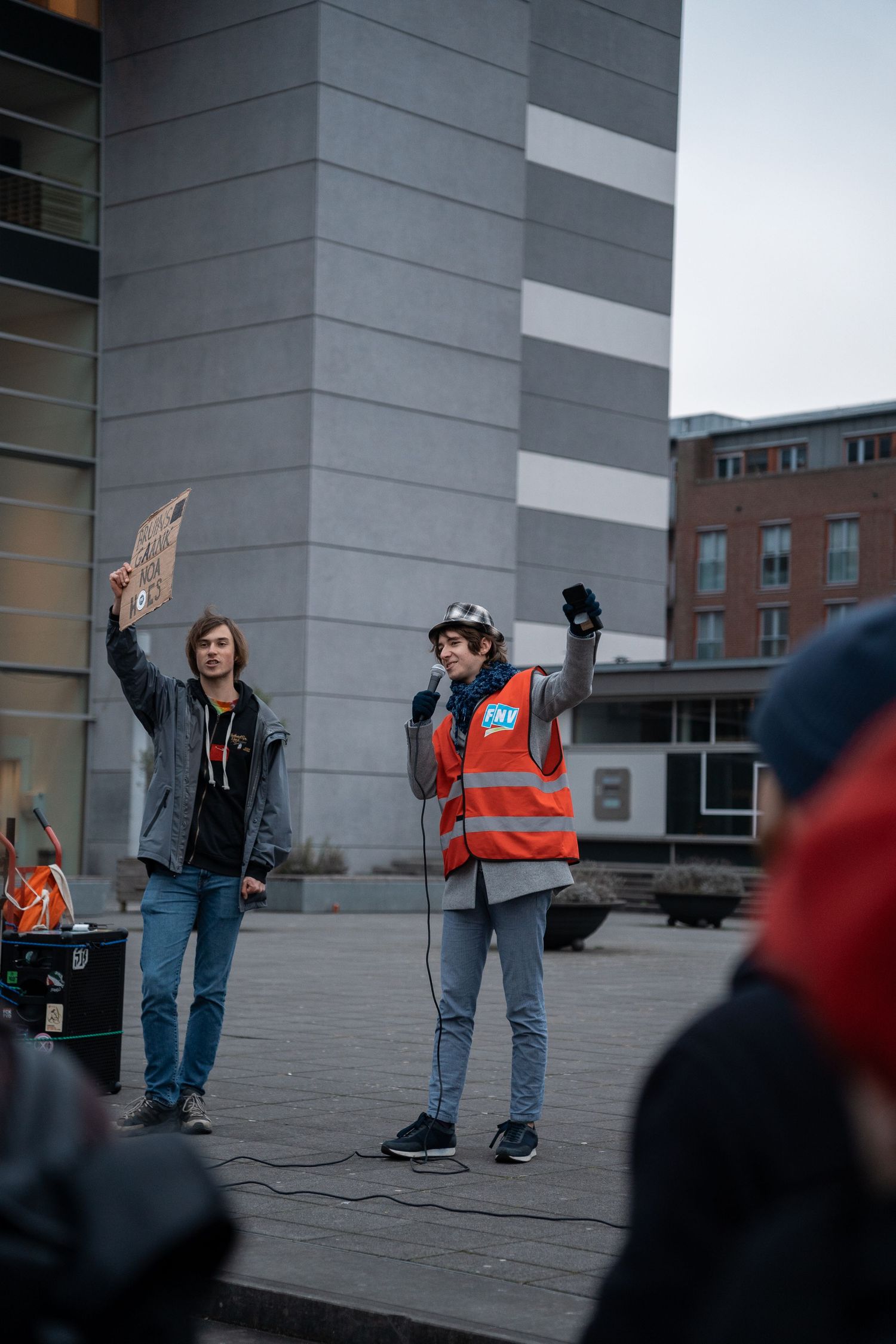 Two speakers address a protest crowd, one wearing an orange safety vest holding a microphone.