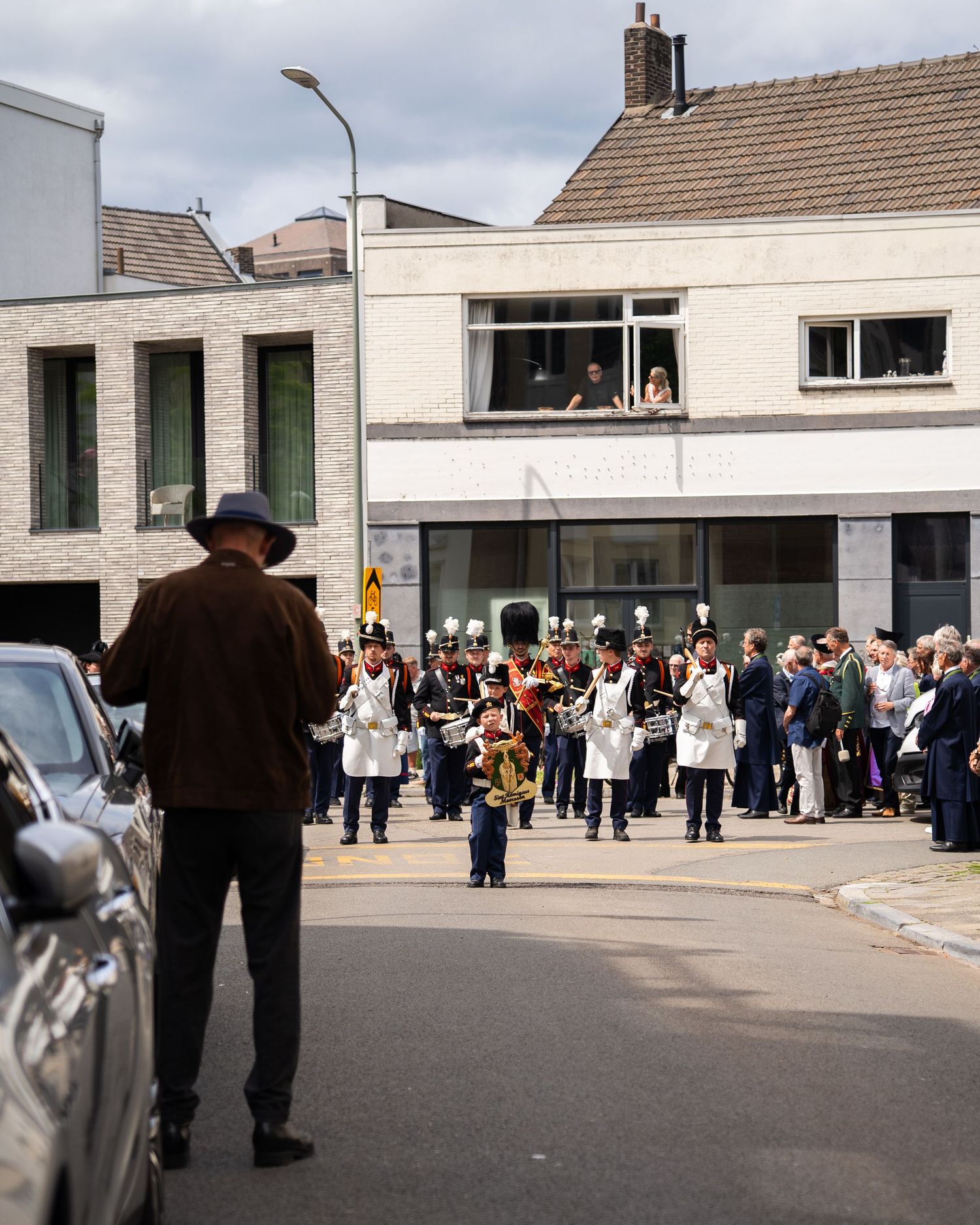 Marching band in white uniforms performs in a street parade while spectators watch from buildings.