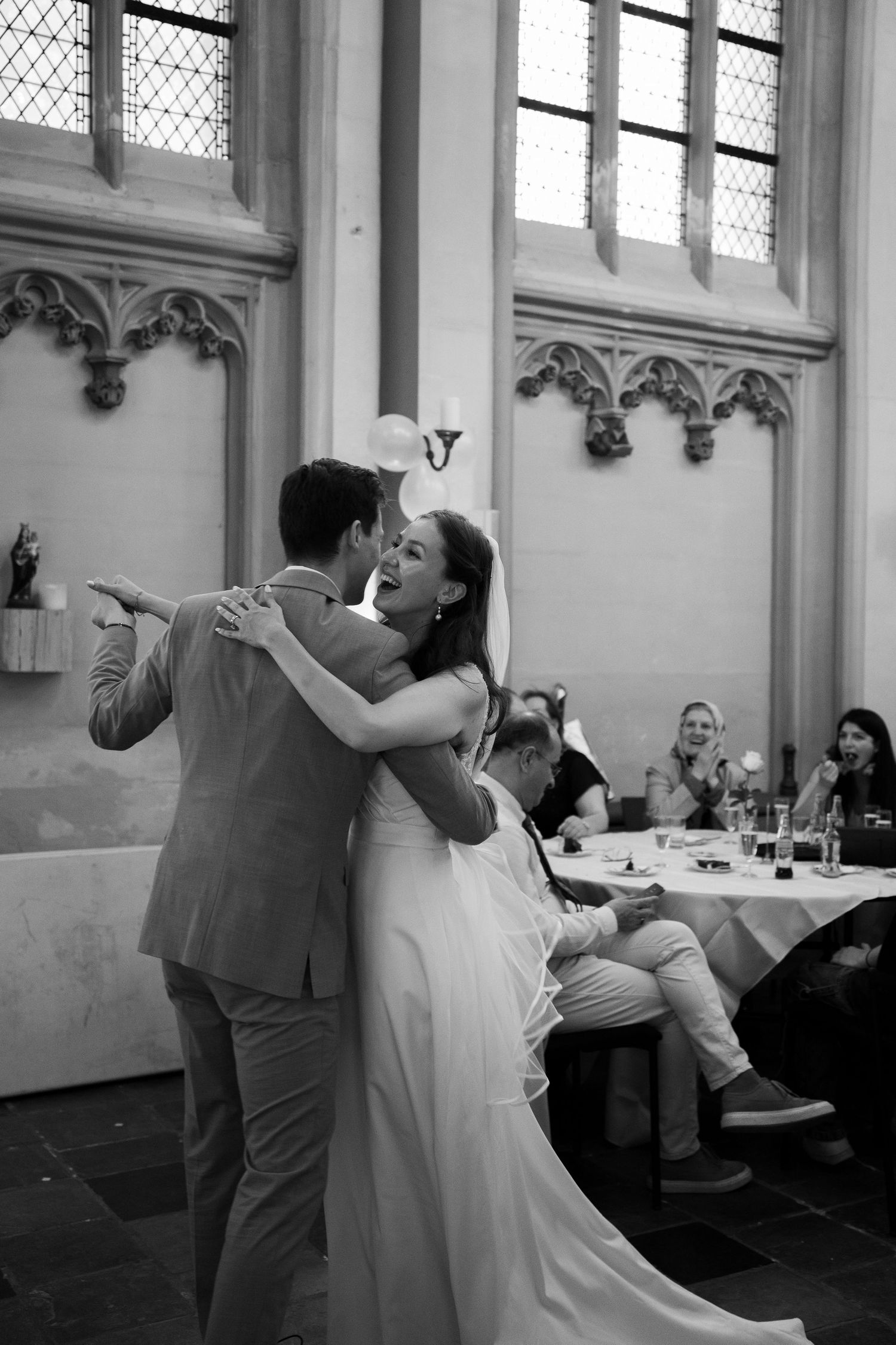 Black and white photo of newlyweds sharing their first dance in a Gothic hall with guests watching.