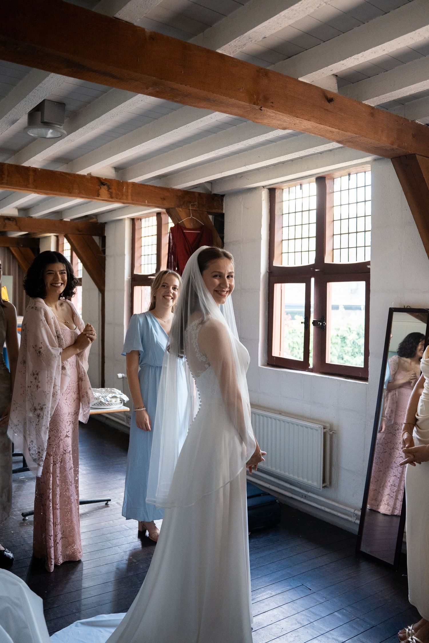 Bride in white gown and veil smiling with bridesmaids in a rustic wooden room.