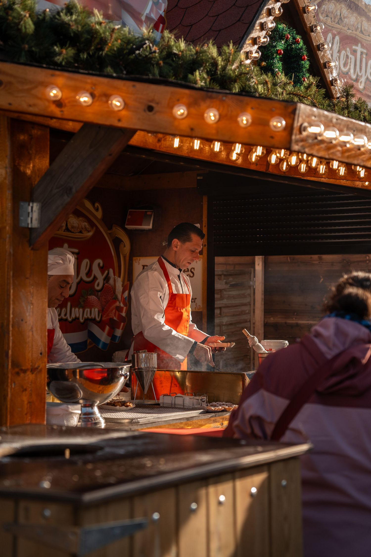 Chef in an orange apron serving food at a warmly lit wooden Christmas market stall during daytime.