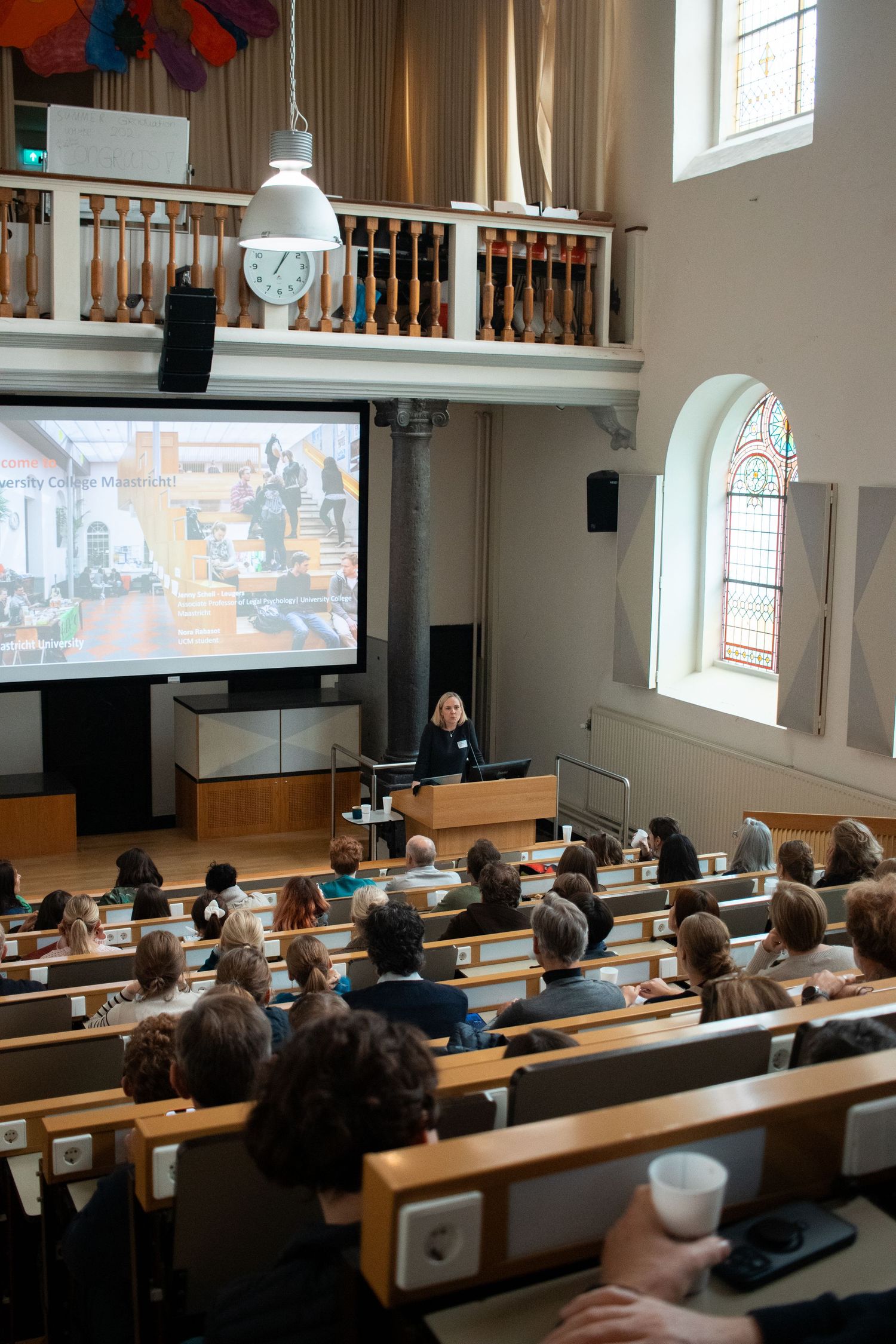 Audience listens to a female speaker presenting on a large screen in a historic lecture hall.