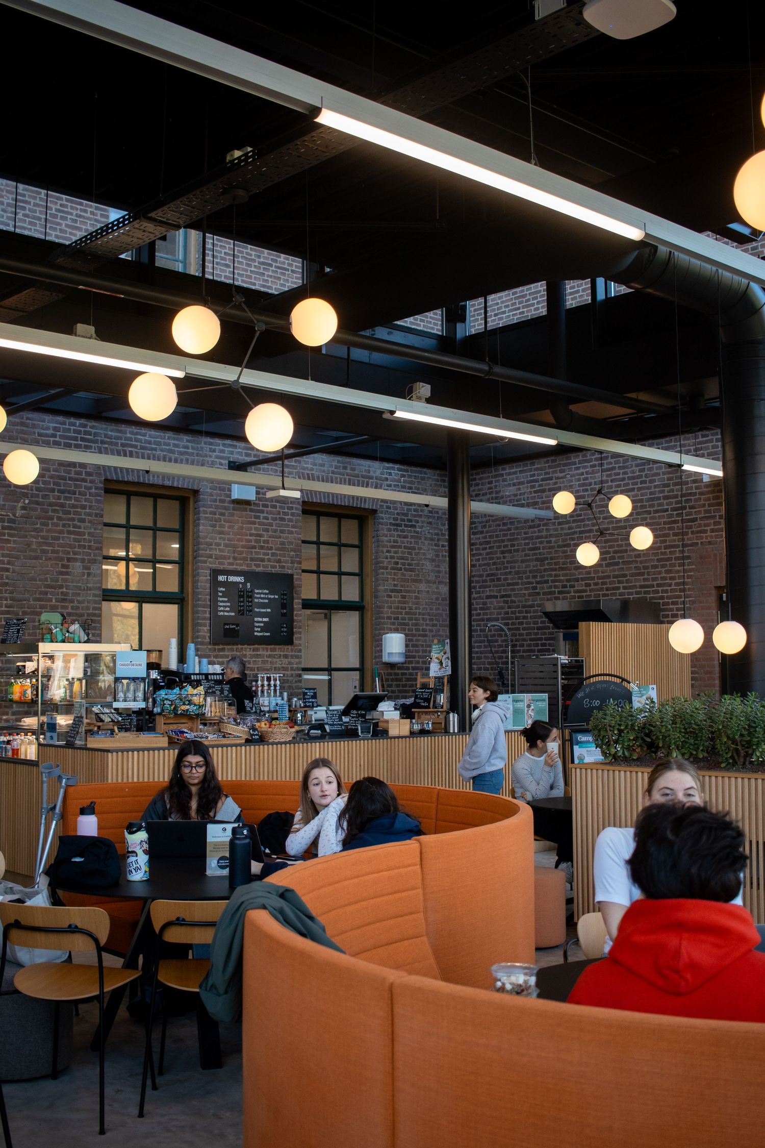 Students relax in orange booth seating at a modern industrial-style university campus cafe.