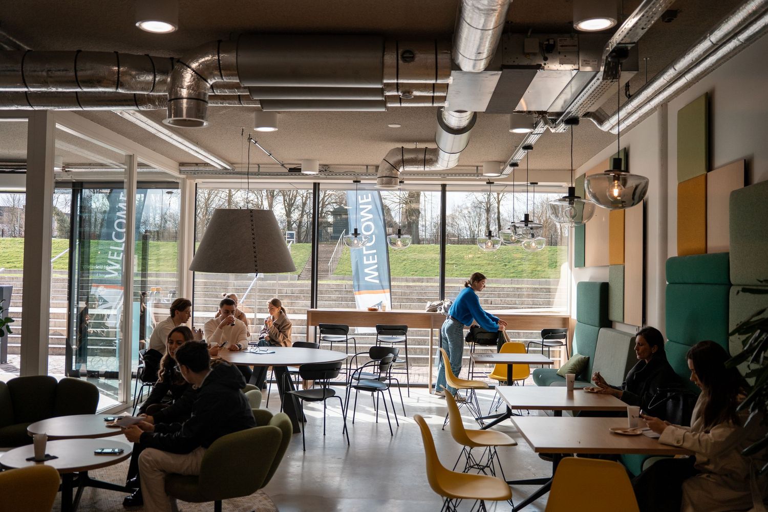 Students relaxing and studying in modern university cafe with industrial ceiling and colorful furniture.