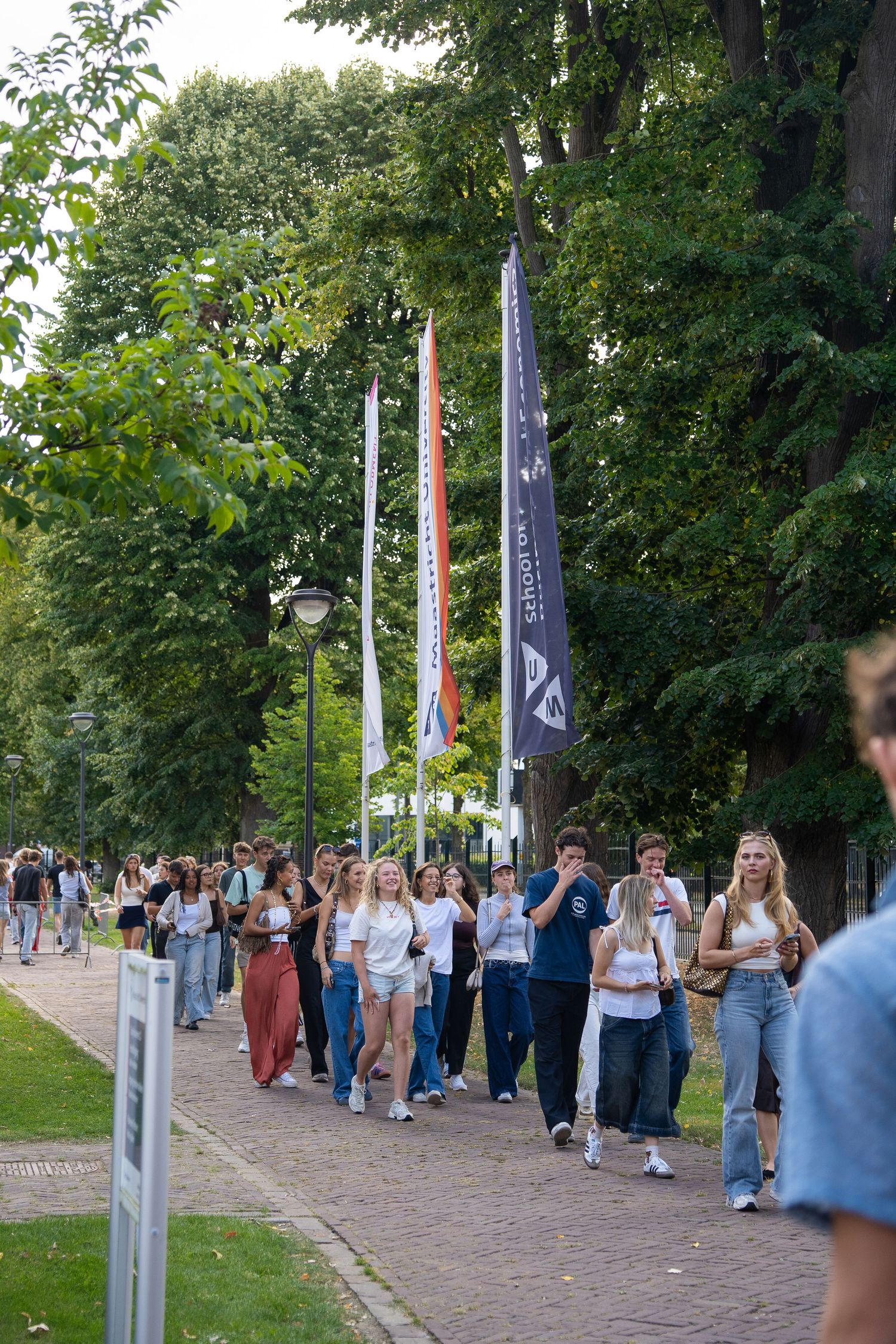Students walk along a tree-lined path carrying bags during a university orientation event with tall flags.