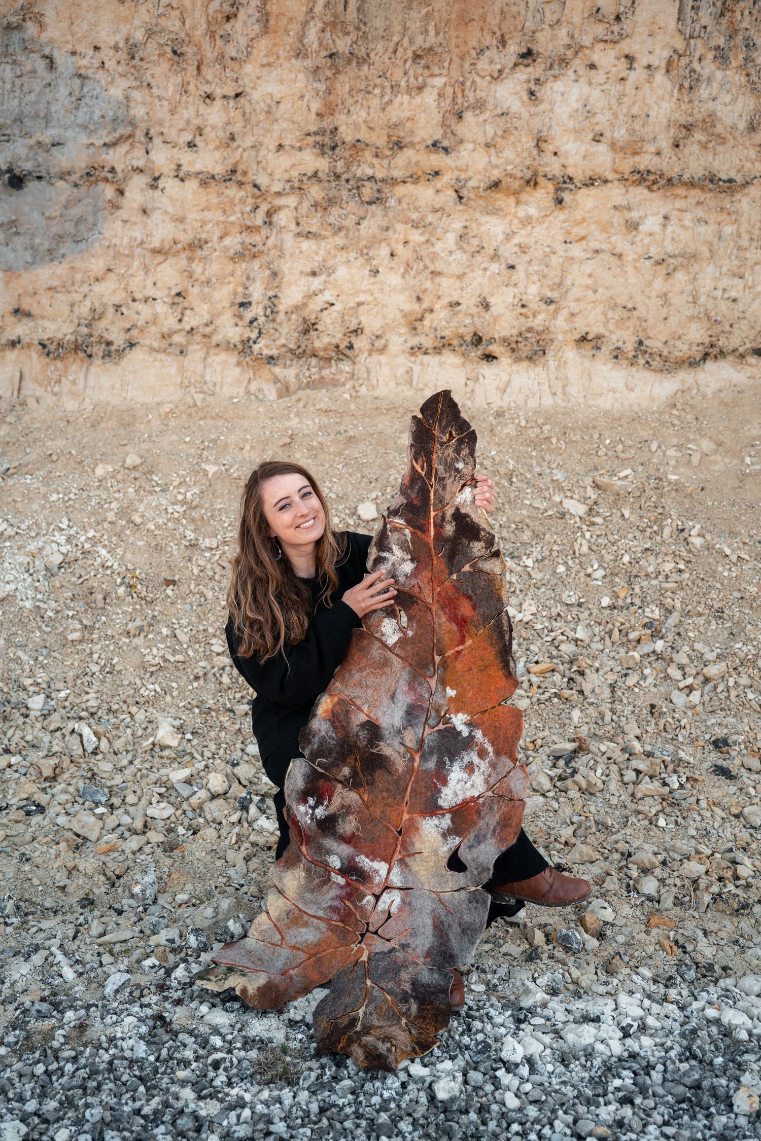 Smiling woman in black crouching beside a large rust-toned felted leaf sculpture on rocky ground.