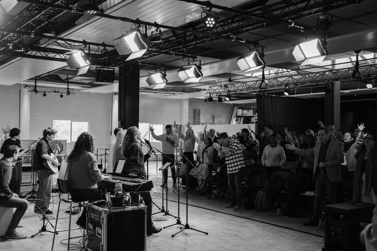 Black and white photo of a live worship band performing on stage before a crowd of raised hands in a church setting.