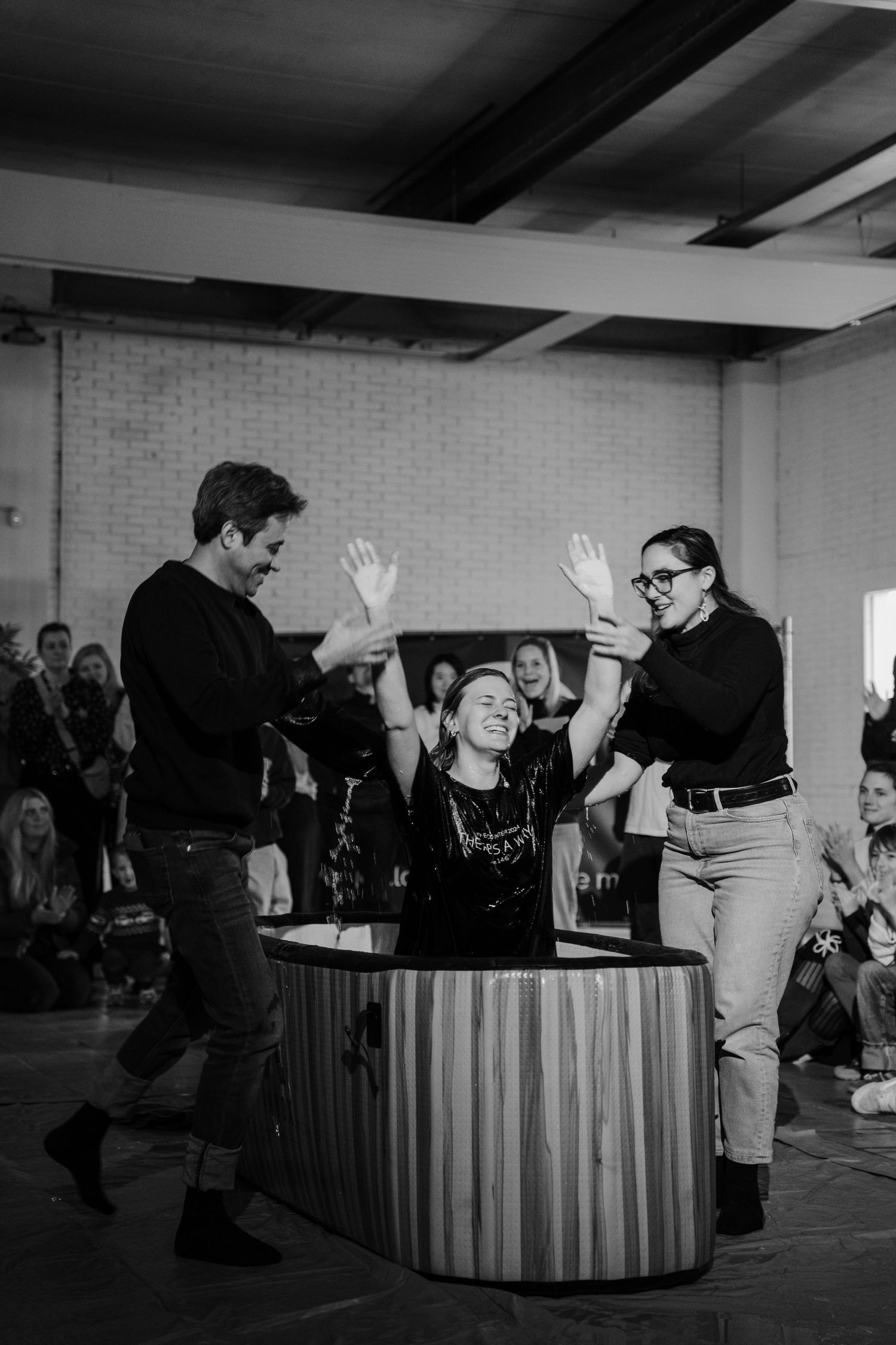 Black and white photo of a joyful woman raising her hands during a baptism ceremony with supporters beside her.