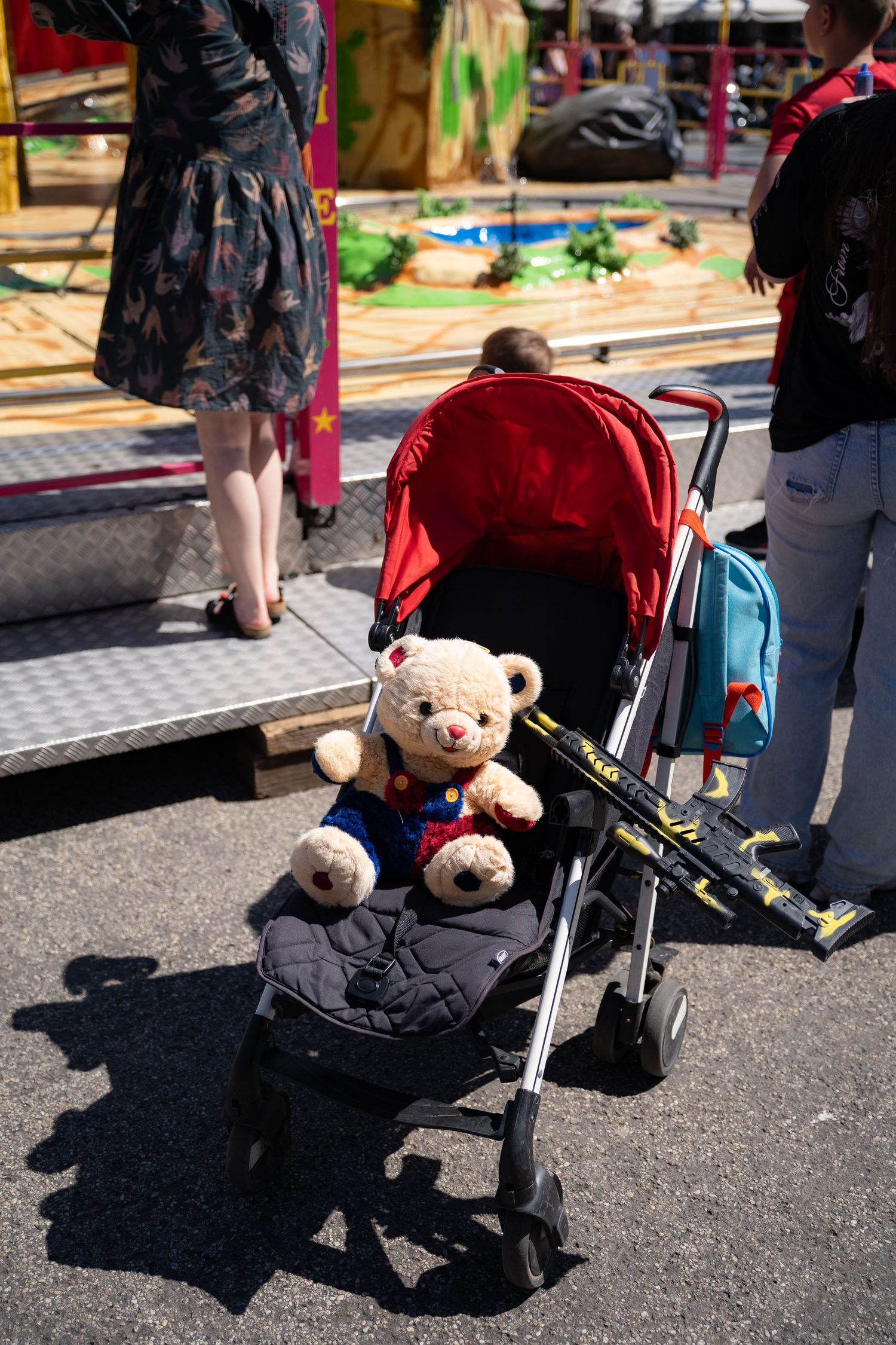 A teddy bear stuffed animal sits in a red baby stroller at a colorful outdoor carnival fairground.