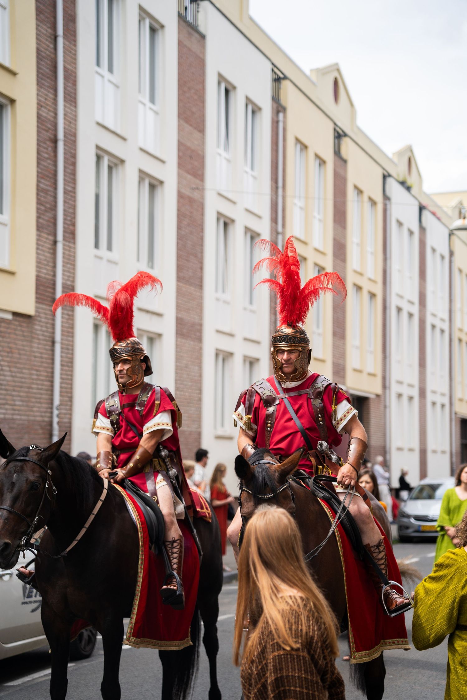 Two riders in Roman soldier costumes with red feathered helmets on horseback in a street parade.